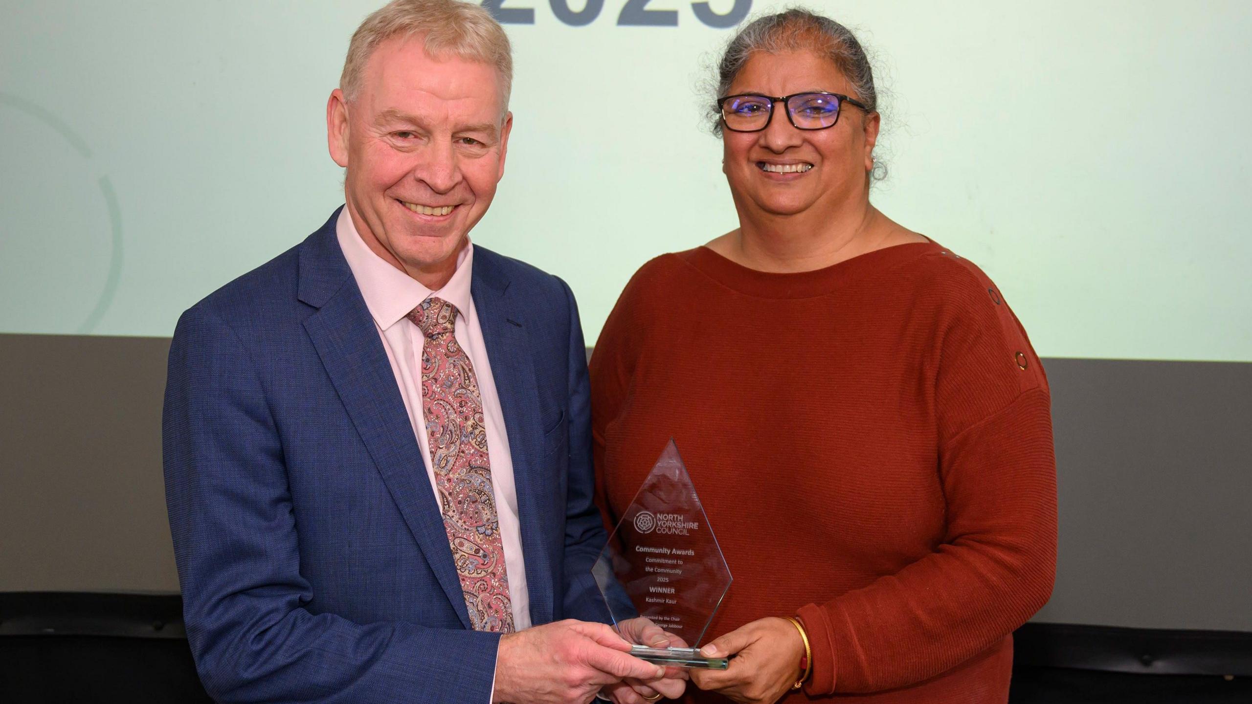 Ms Kaur stands side by side with North Yorkshire's Council's CEO Richard Flinton, receiving her award at North Yorkshire’s Community Awards. They both hold a side of a diamond-shaped glass award in their hands. Mr Flinton has short white hair and wears a pink shirt, pink patterned tie and a blue suit jacket. Mr Kaur has grey hair worn up, and wears black-framed glasses and a red long sleeved top.