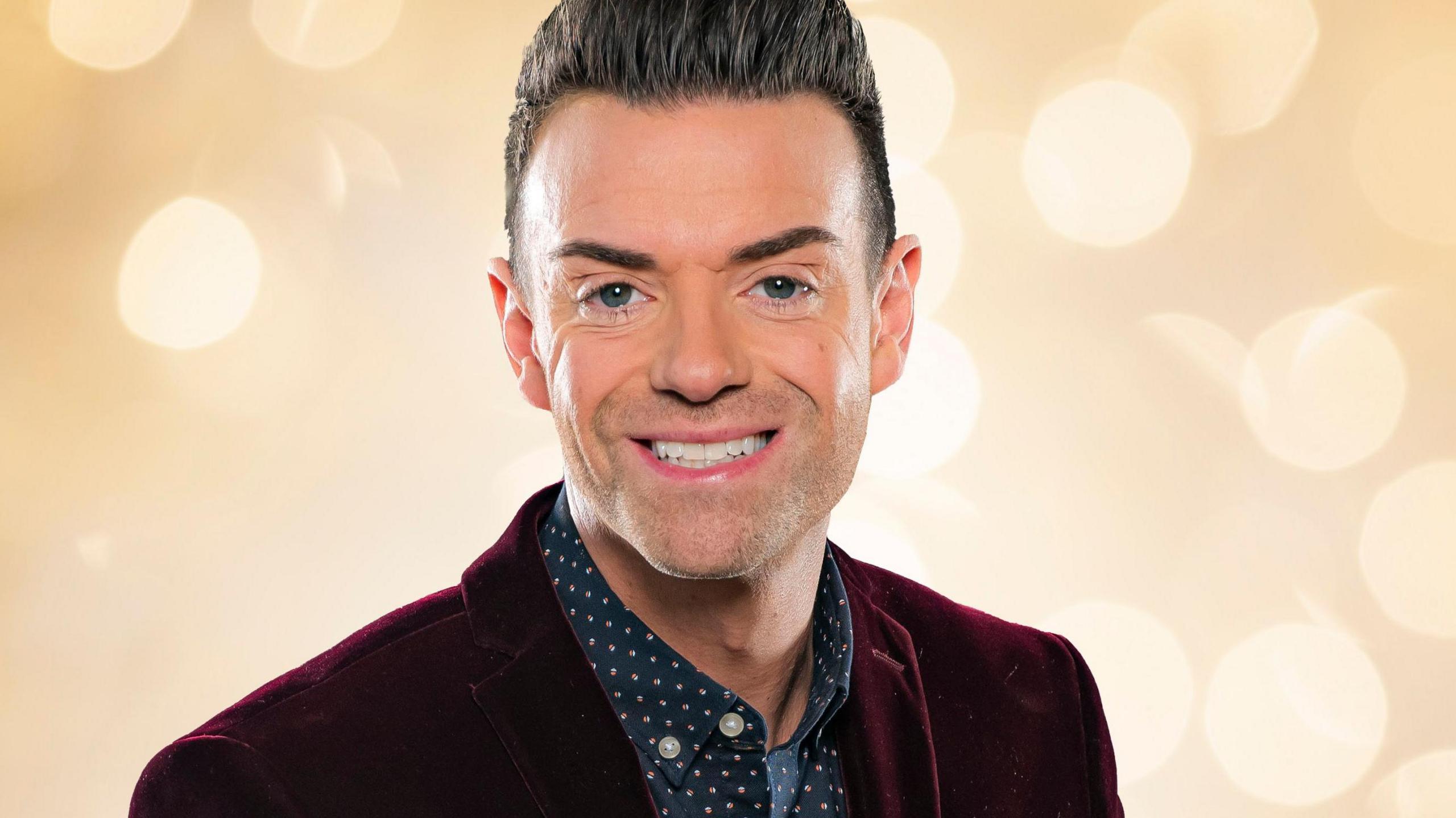 Man with slicked back brown hair smiles directly at the camera in a publicity shot. 