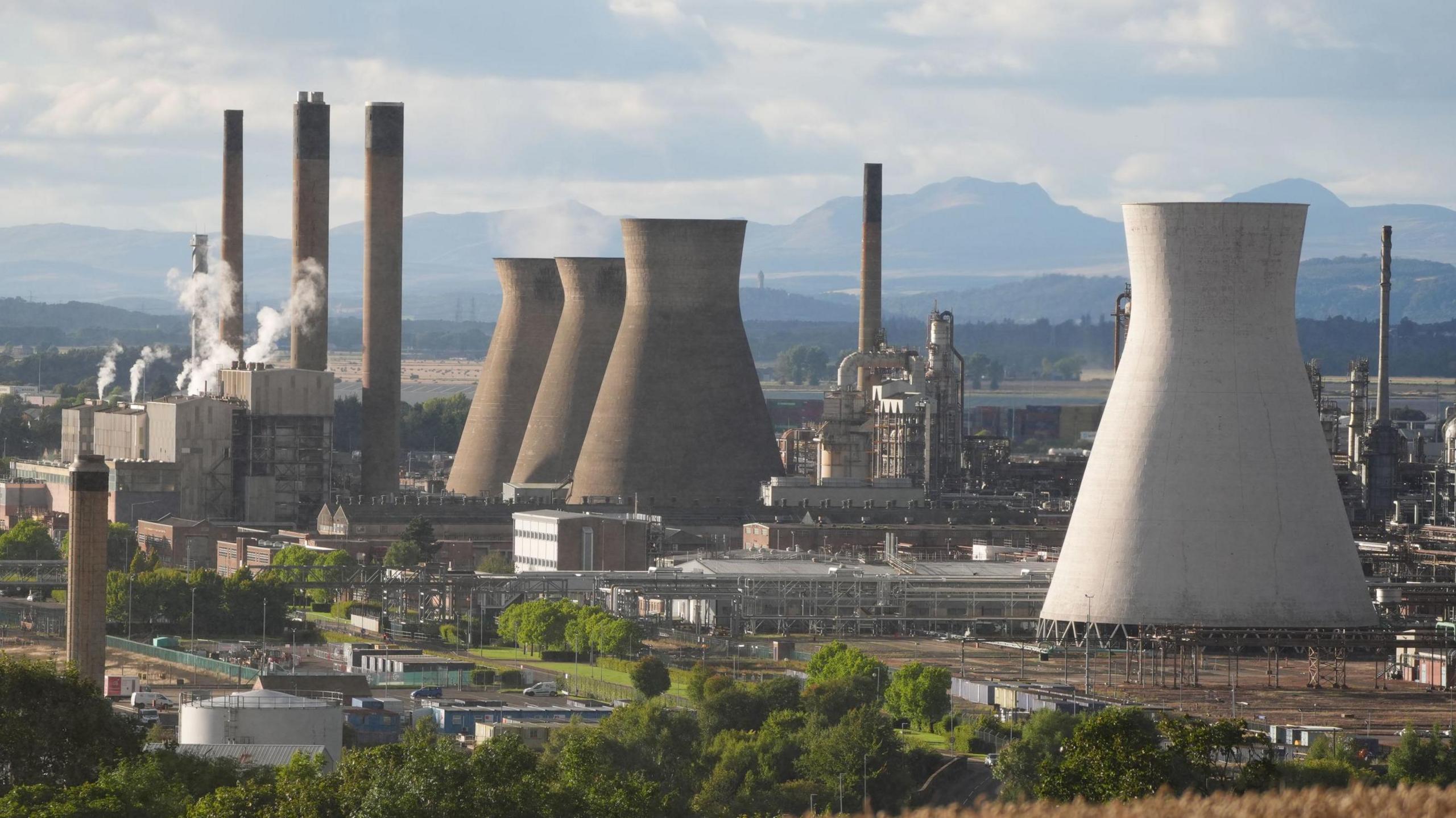 A view of the Grangemouth petrochemical plant in Grangemouth. A large white tower is in the foreground, with three darker ones further away. Smoke billows from the chimneys f various buildings in the heavily industrial site