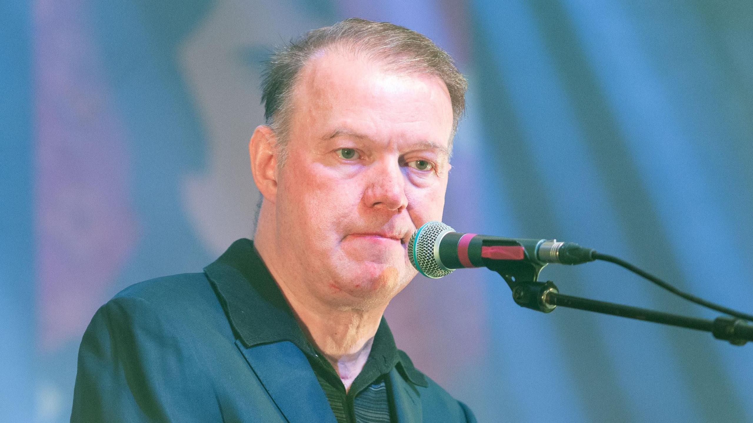 Edwyn Collins sitting on stage at a microphone - he is wearing a blue jacket and a green T-shirt and has thinning brown hair