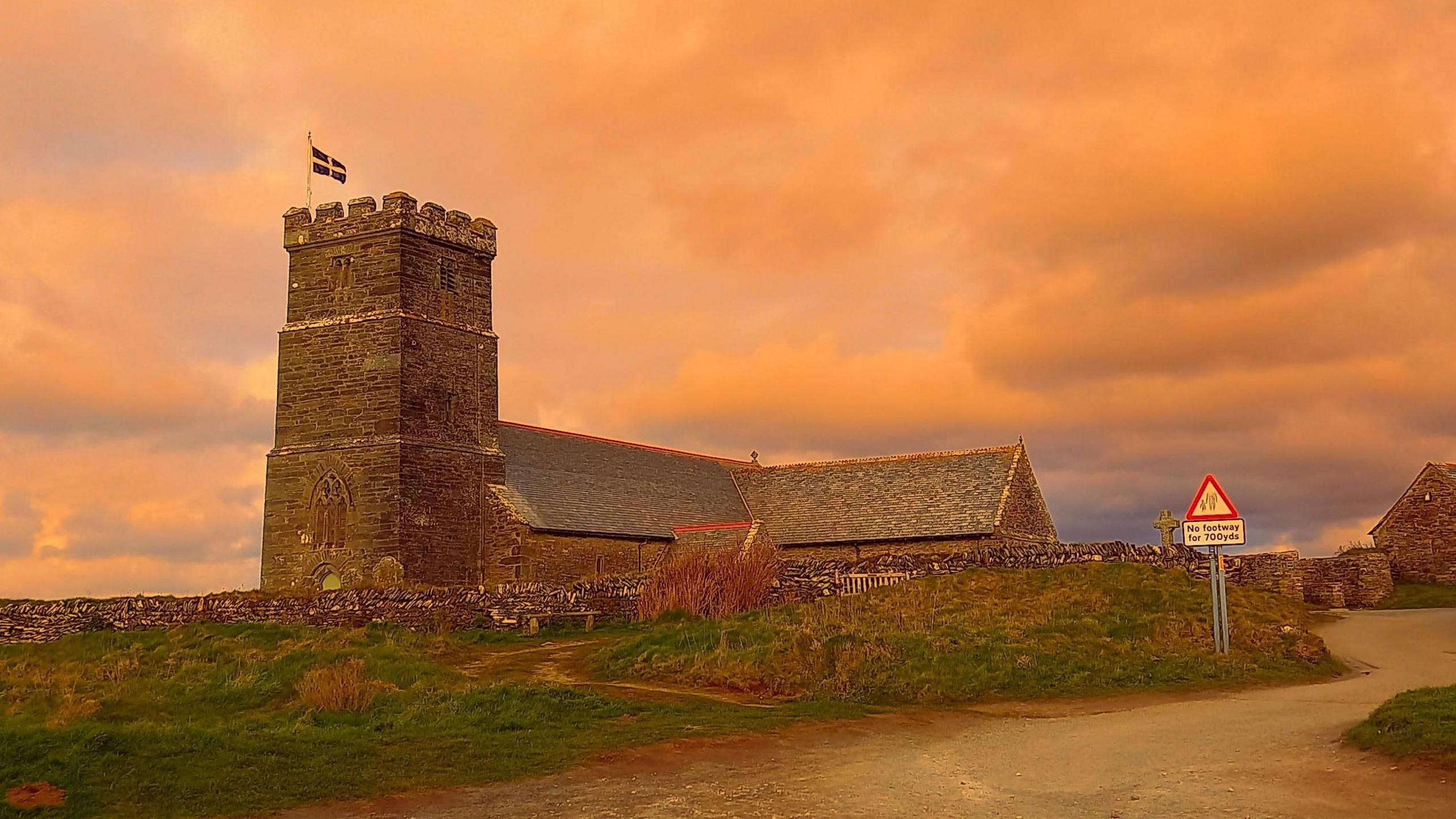 A church in Tintagel in Cornwall. The sky has an orange hue. A Cornish flag is flying above the church's spire.