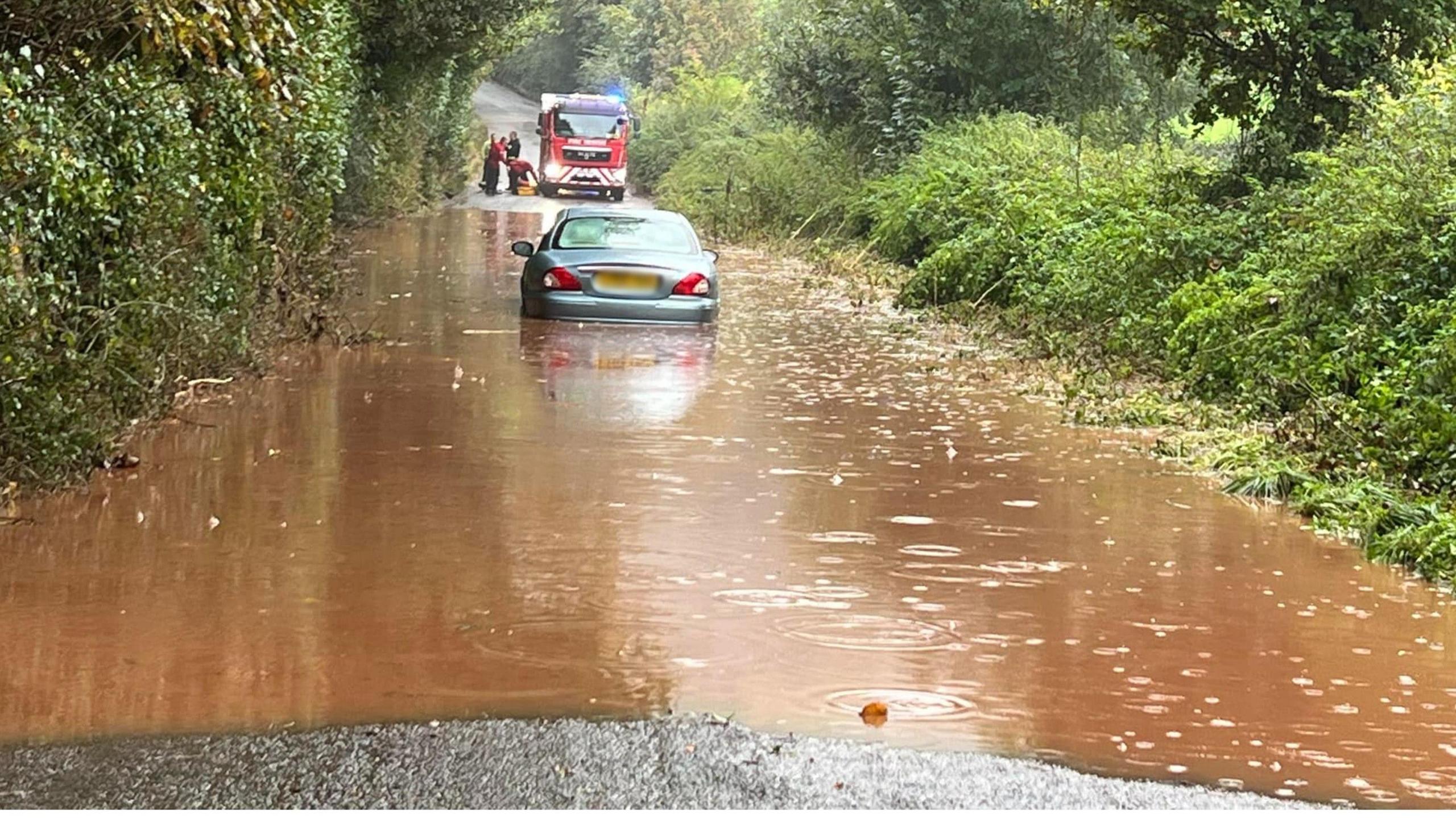 A silver vehicle in the middle of a flooded road heading into Crediton from Sandford. The wheels of the car are under the brown coloured flood water. In the background is a red fire engine with its blue lights on. To the right of the fire engine are crews. 