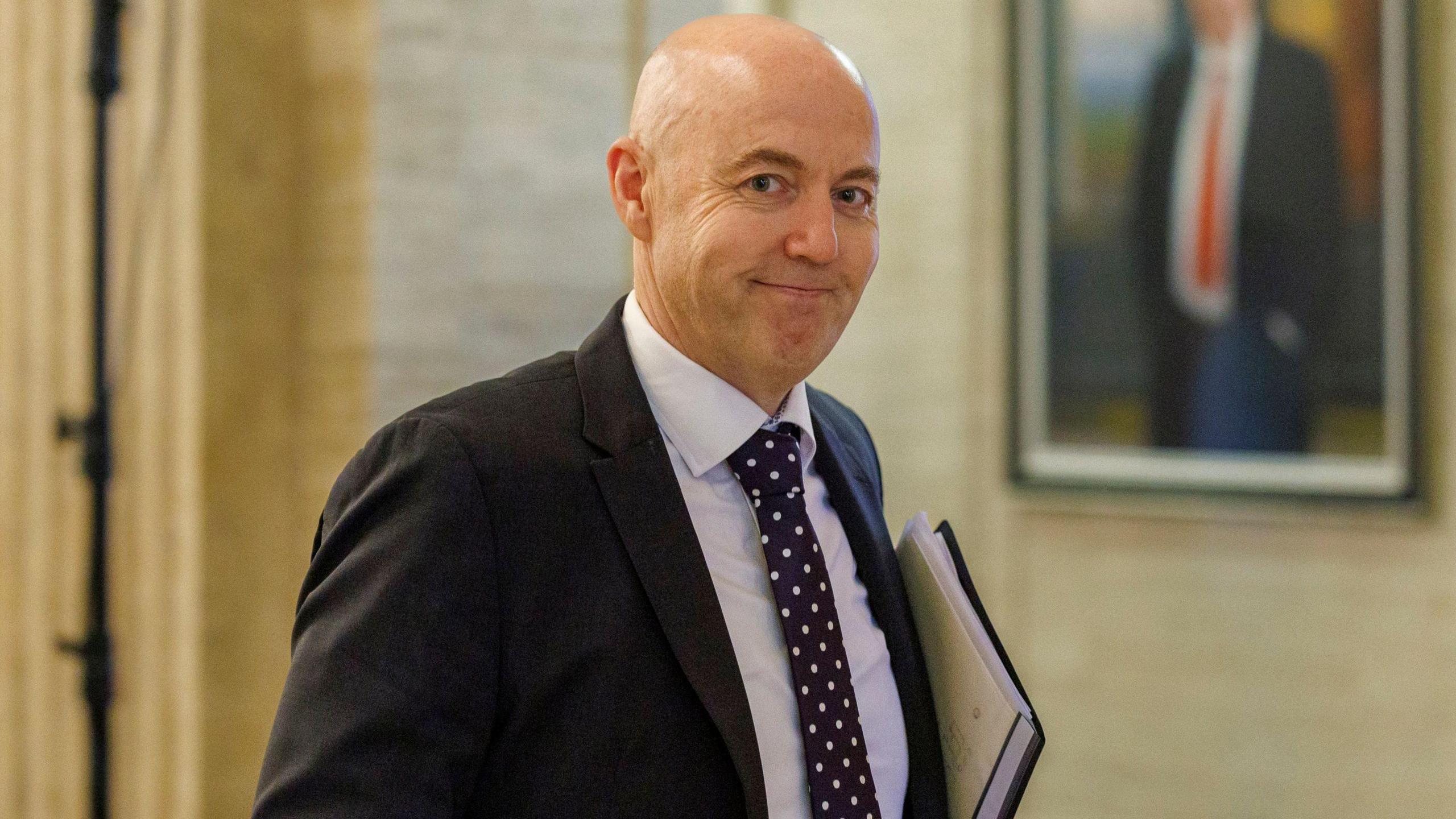 Jon Burrows is smiling directly at the camera. He is wearing a black suit and white shirt. His navy tie has white sports on it. He has a bald head and is carrying a folder with a A4 paper in it under its arm. 