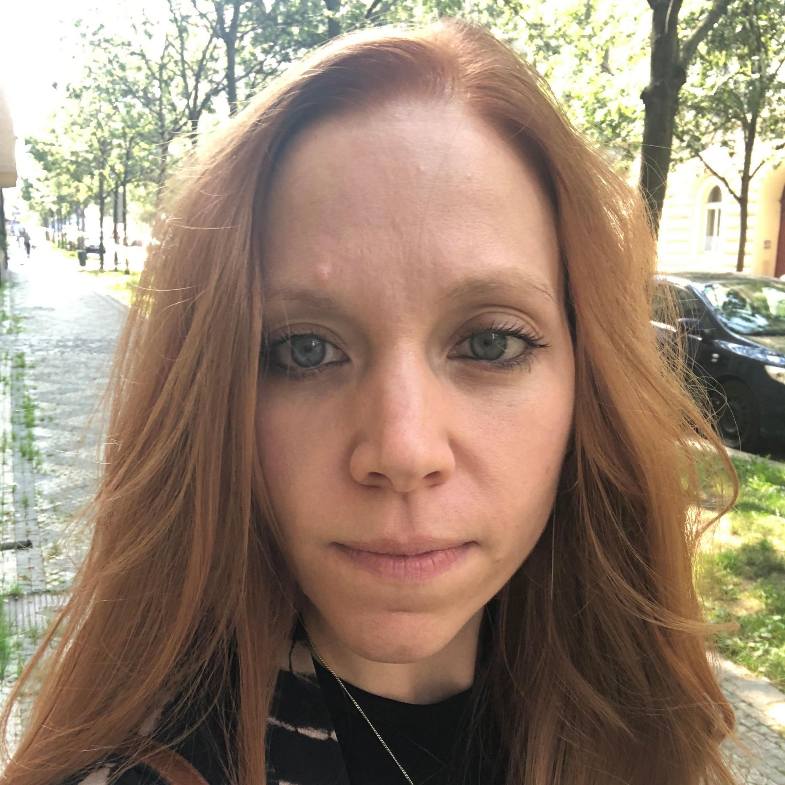 A close-up image of a red-haired woman. She is wearing a black top and is standing in a tree-lined street.