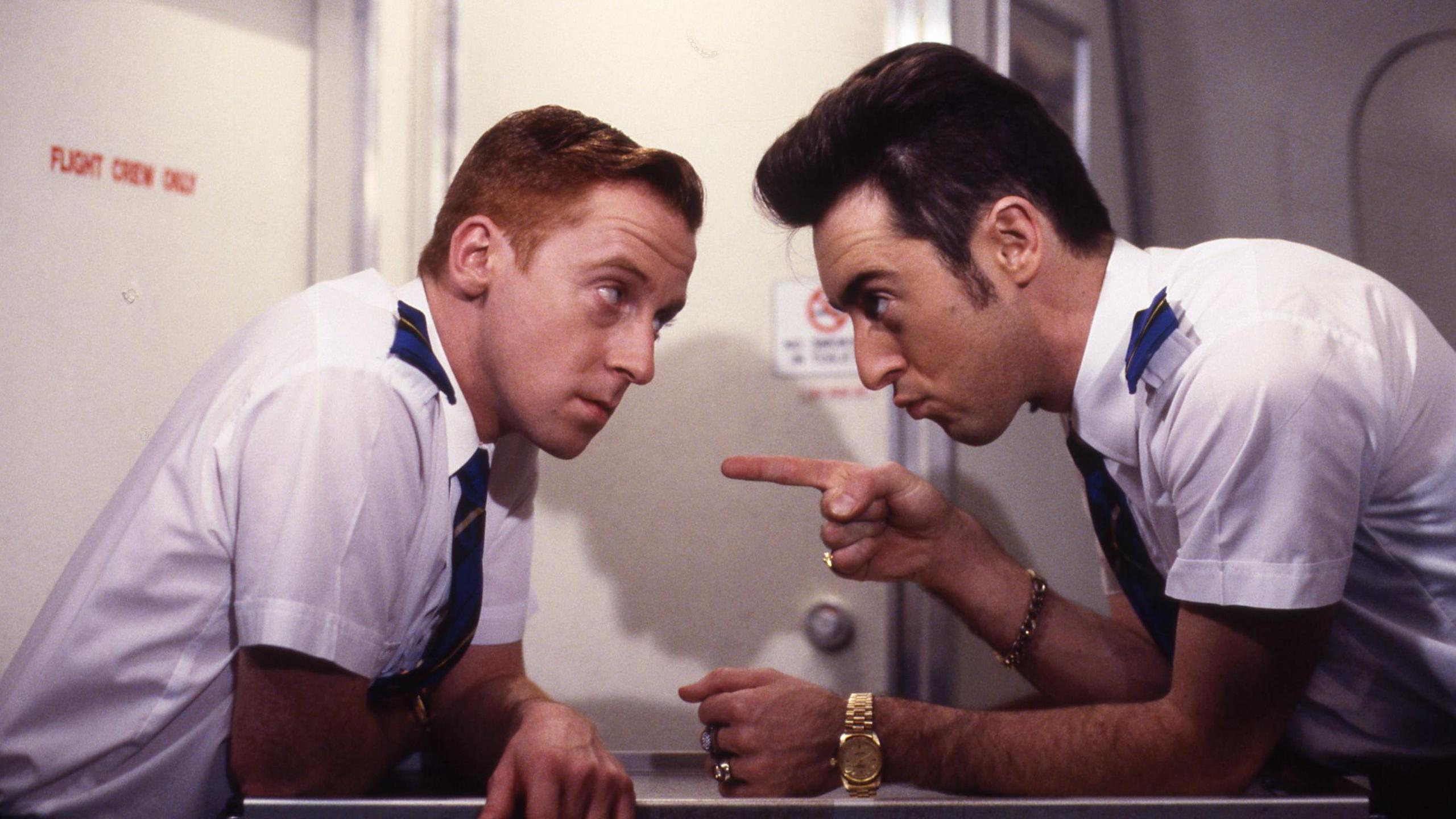 Alan and Forbes Masson in character on sitcom The High Life - both are dressed as airline stewards, with white shirts and blue ties. They are seated at a table staring at each other, with Cumming pointing a finger at Masson, as if they are arguing.