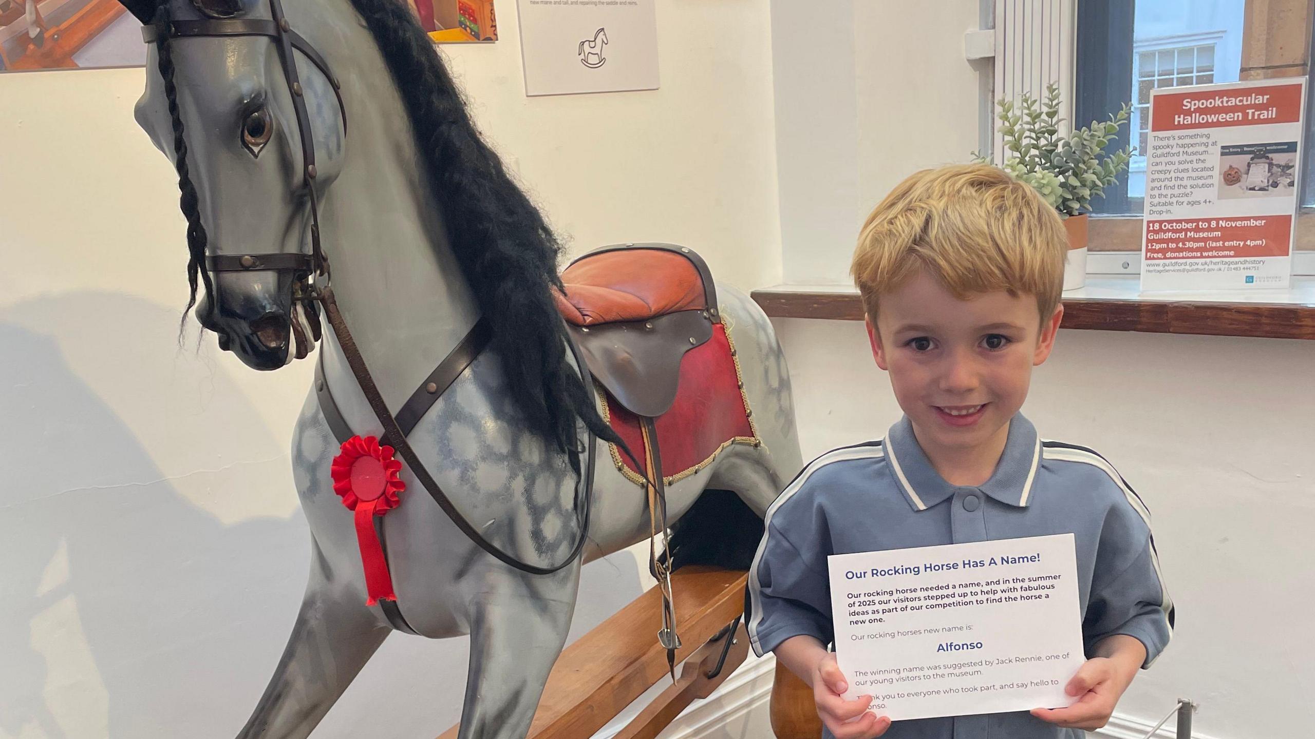 A boy with blonde hair smiling at the camera while holding a white certificate. He is stood beside a large grey rocking horse