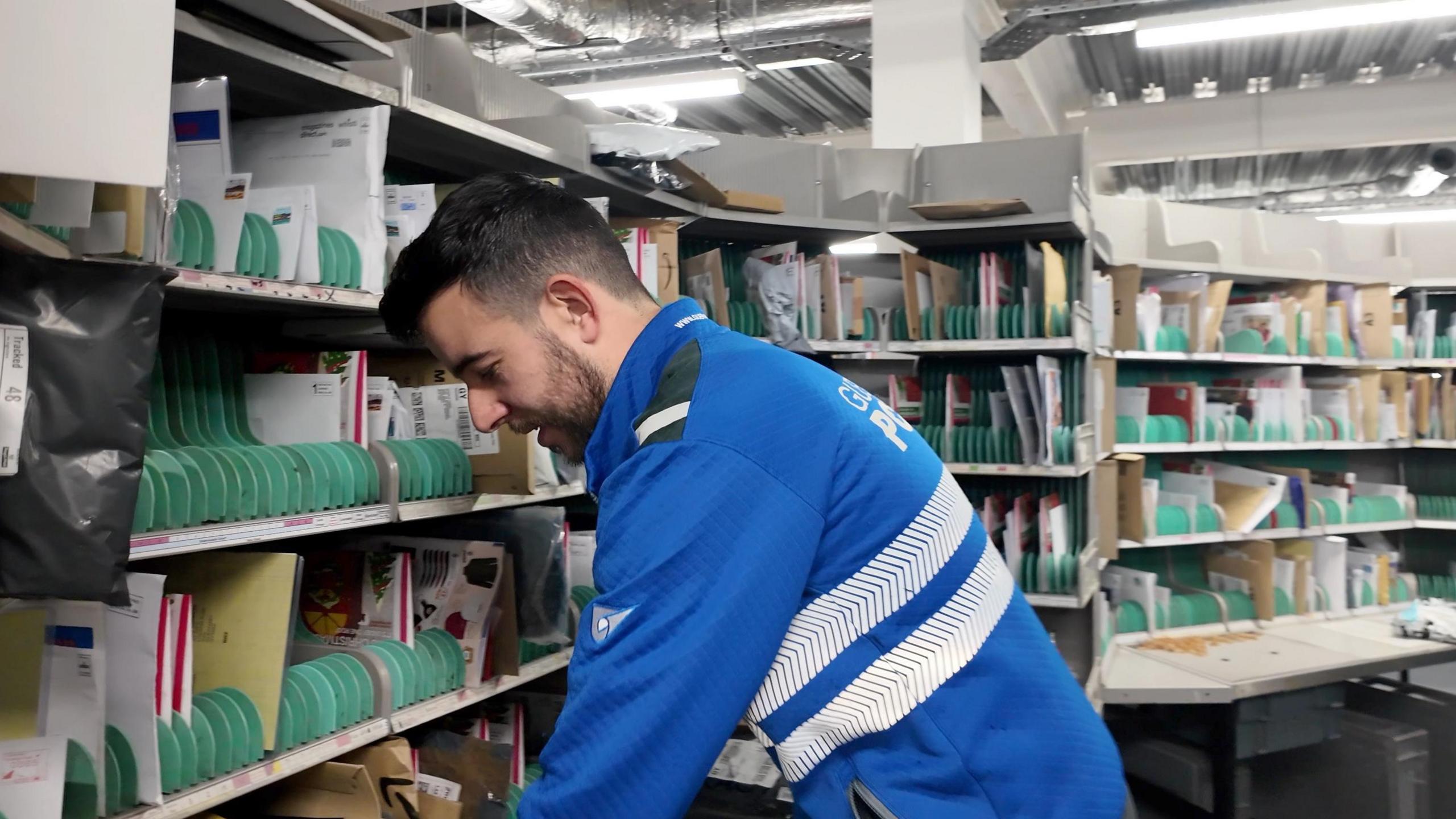 A man with dark hair in a blue Guernsey post jacket sorting letters, surrounded by shelves of letters in a post sorting office.