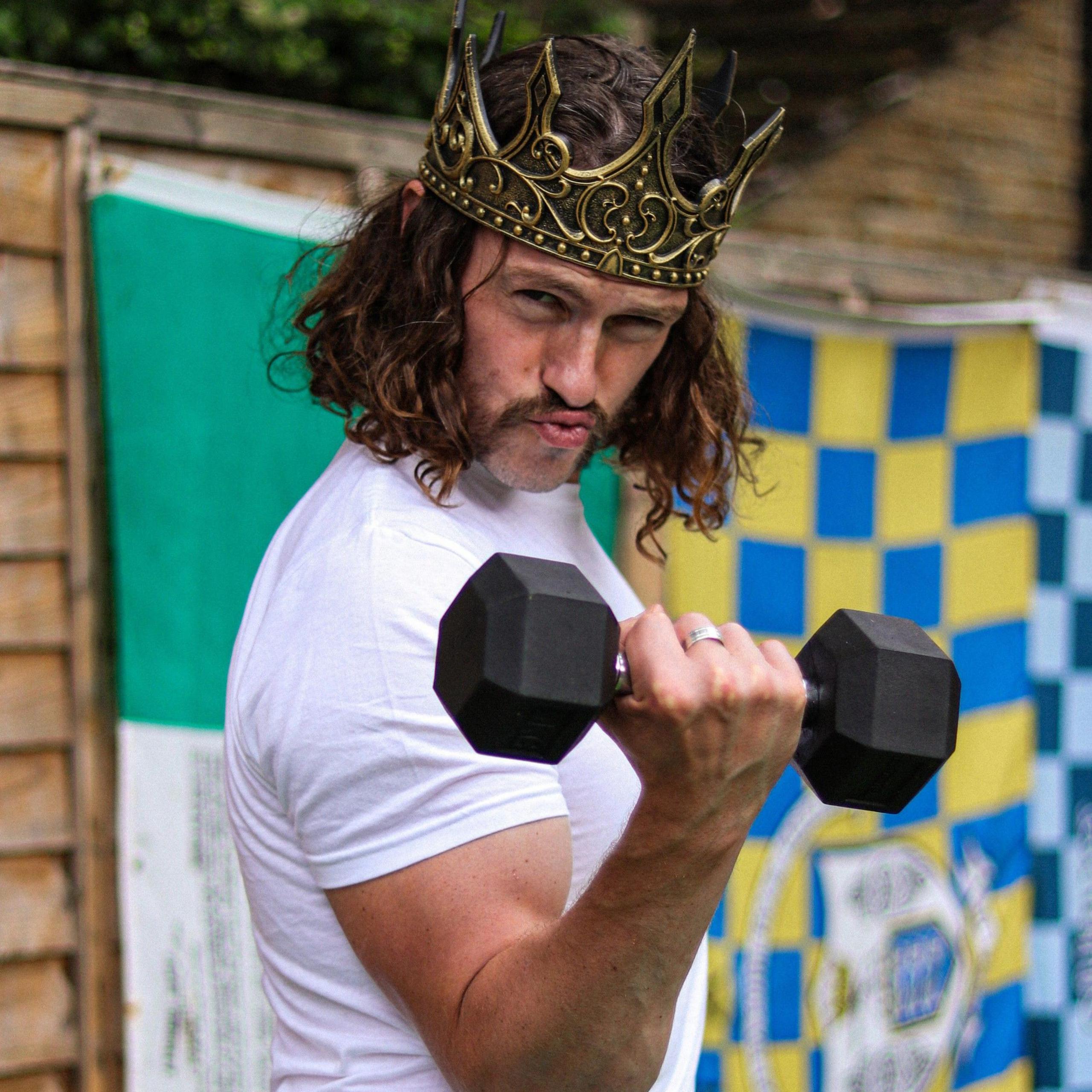 Iain Ward is seen with long hair and a moustache wears a gold crown and white T-shirt while lifting a dumbbell. He poses playfully with pursed lips, with colourful flags hanging in the background.