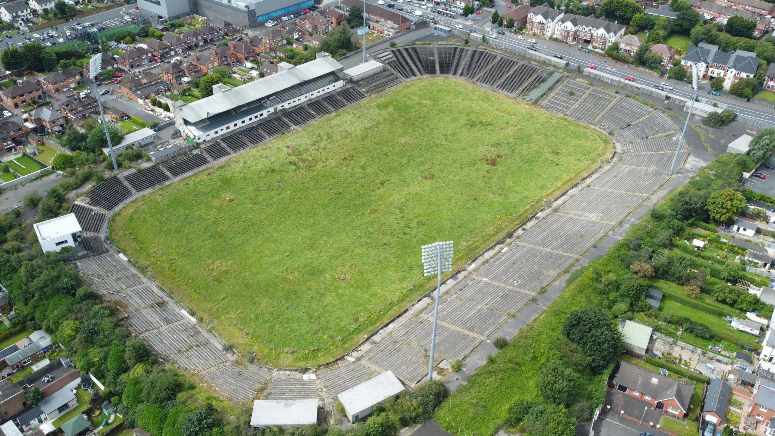 Casement Park with its green worn grass and grey stands, and surrounding area with clutters of houses and main roads 