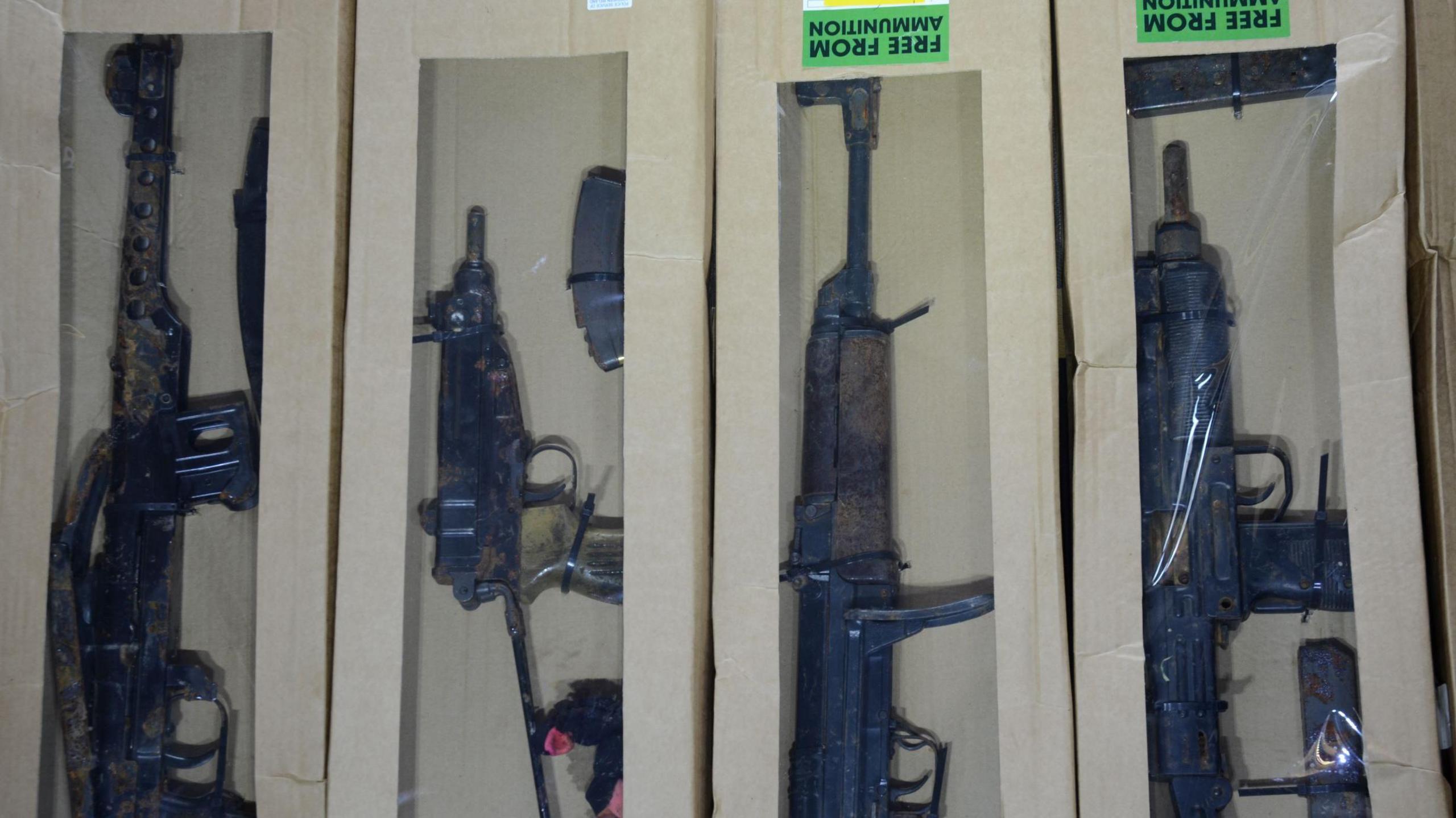 A photo of four of the 11 guns which were hidden by the conspirators in a field for the NCA to find.   The weapons are packed into white boxes with clear plastic covers.  Two of the boxes are marked with green labels which read: "Free from ammunition".