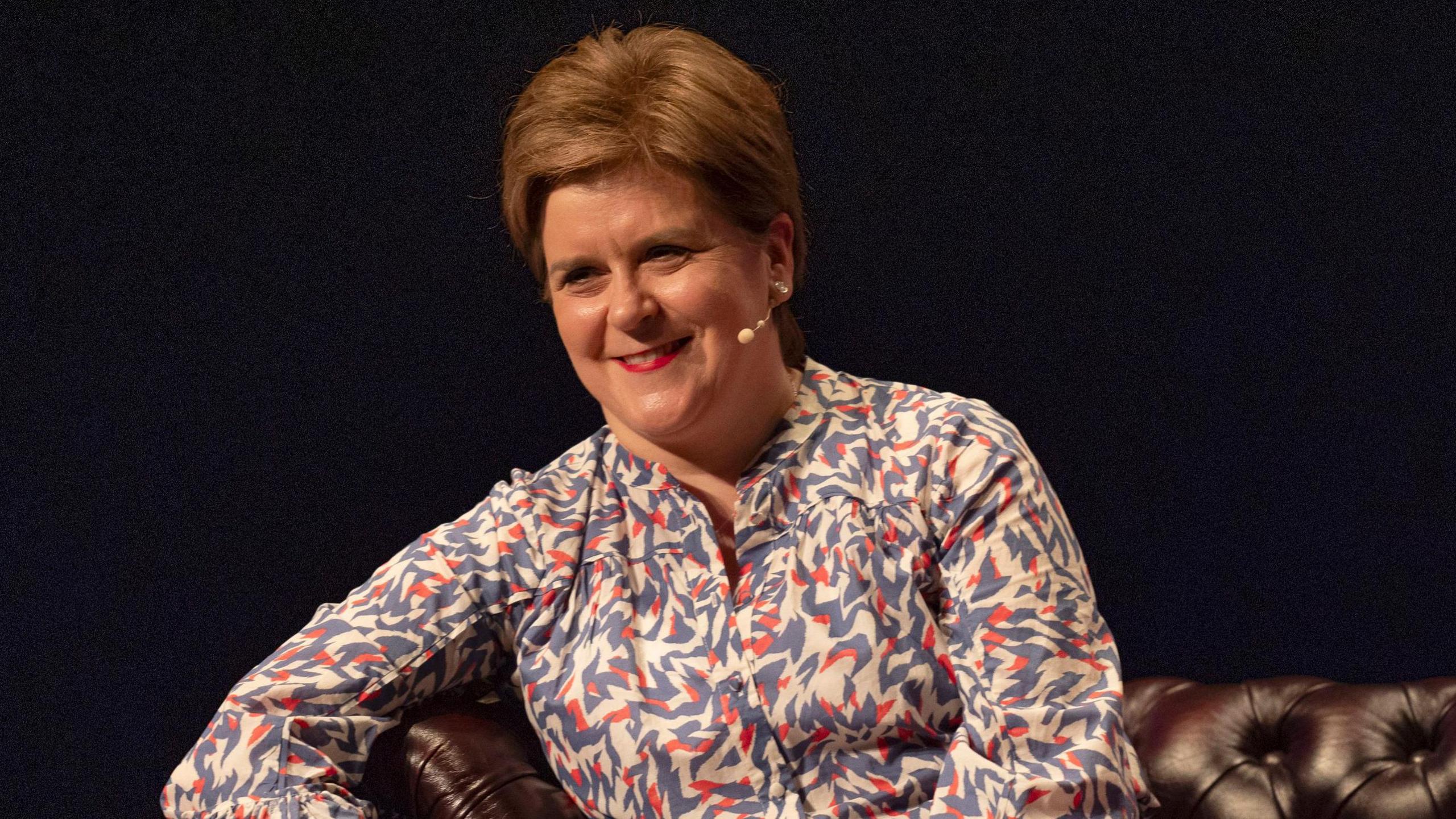 Nicola Sturgeon smiling and looking slightly off camera. She has short, orange hair and red lipstick. She is wearing a multi-coloured shirt and has her right arm slightly raised on the side of a brown sofa