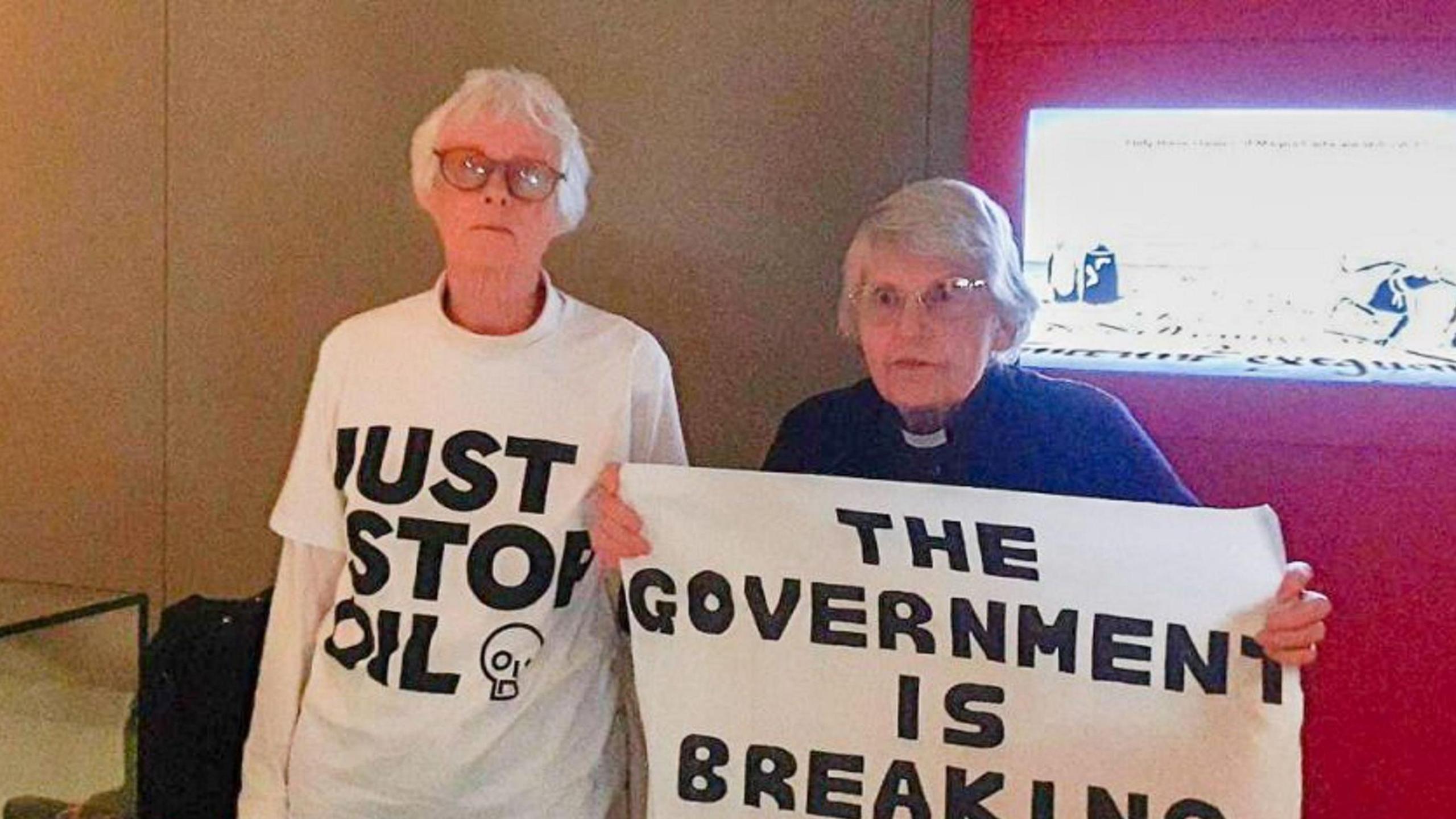 Image of two women, one wearing a JSO t-shirt and the other holding a placard reading "the government is breaking the law"