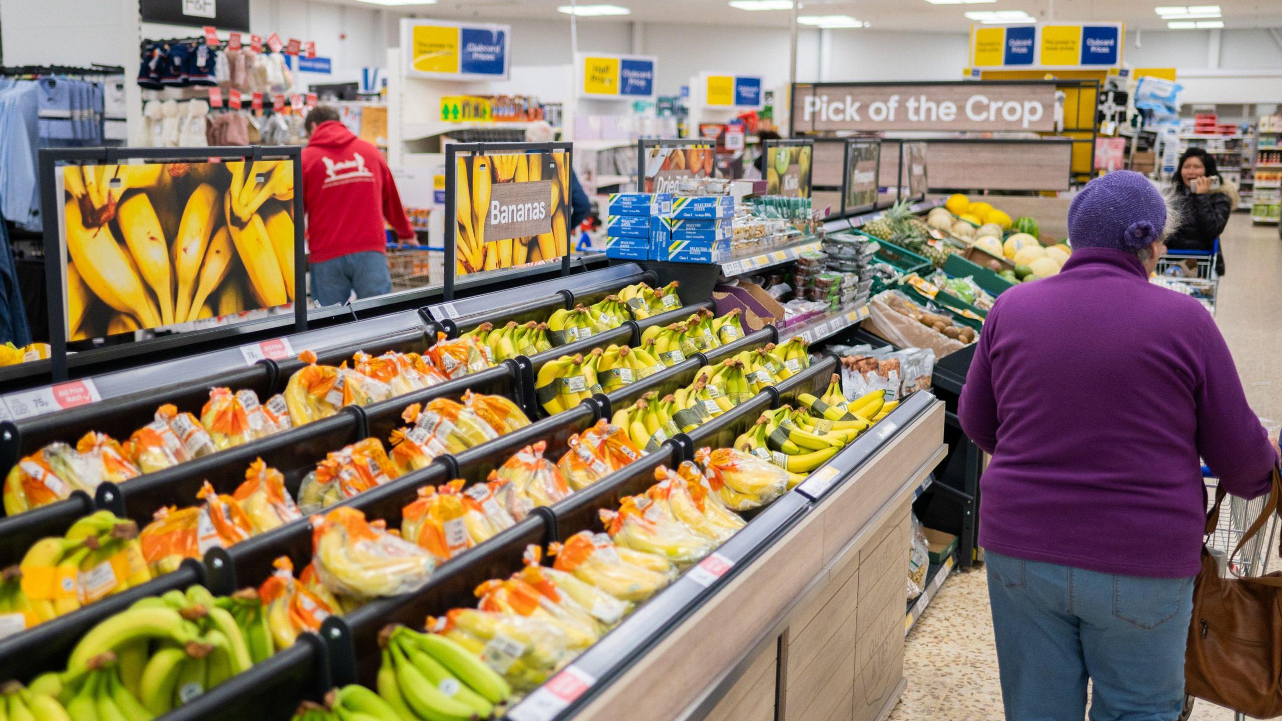 A shopper in a purple sweater pushes a trolley in a Tesco supermarket beside a fresh fruit stand