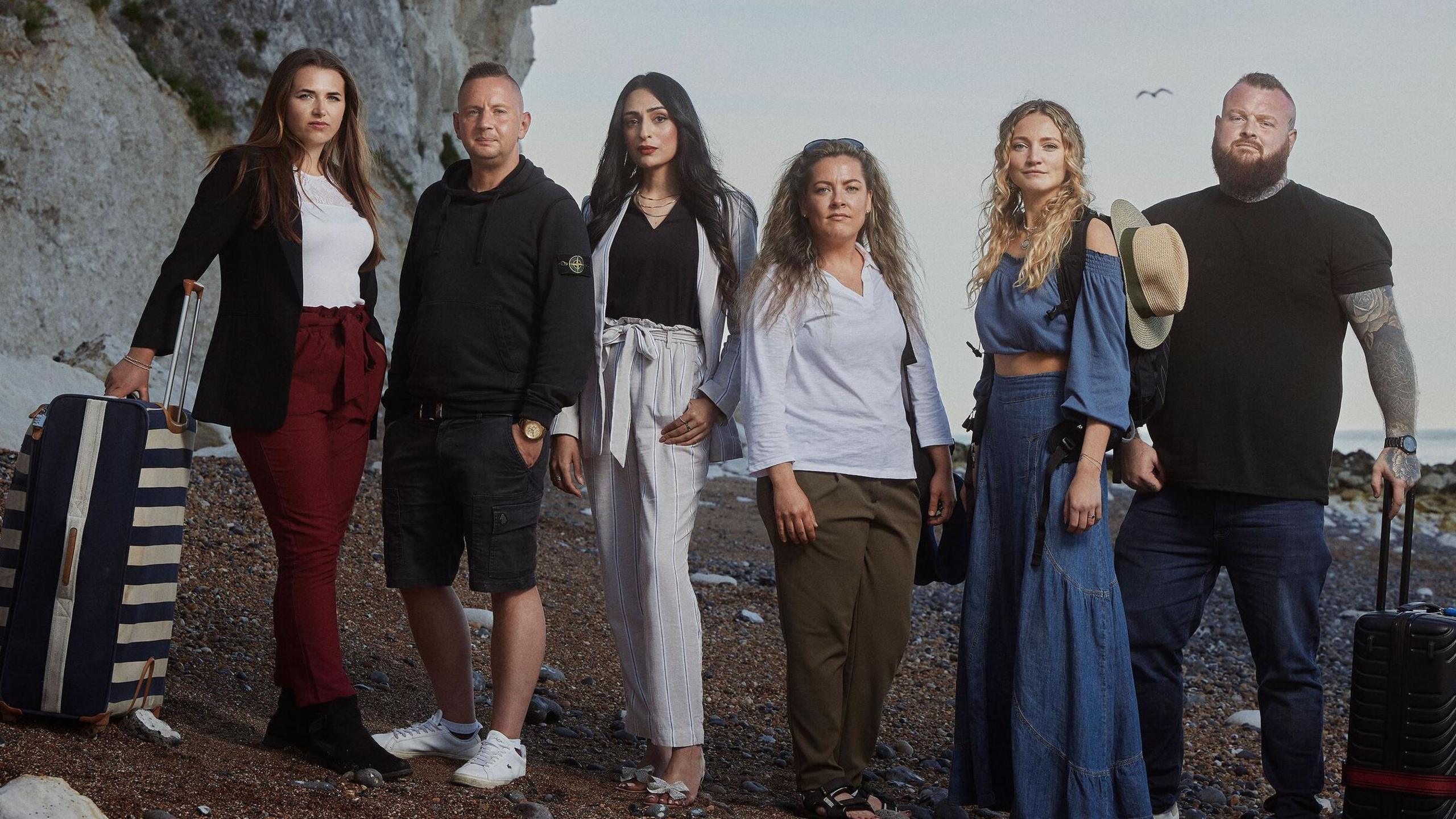 The participants of Go Back To Where You Came From on Dover beach. L-R is Chloe, Dave, Bushra, Jess, Mathilda and Nathan. They all have suitcases as if prepared to go on holiday and stand on the shingle in front of the white cliffs. 