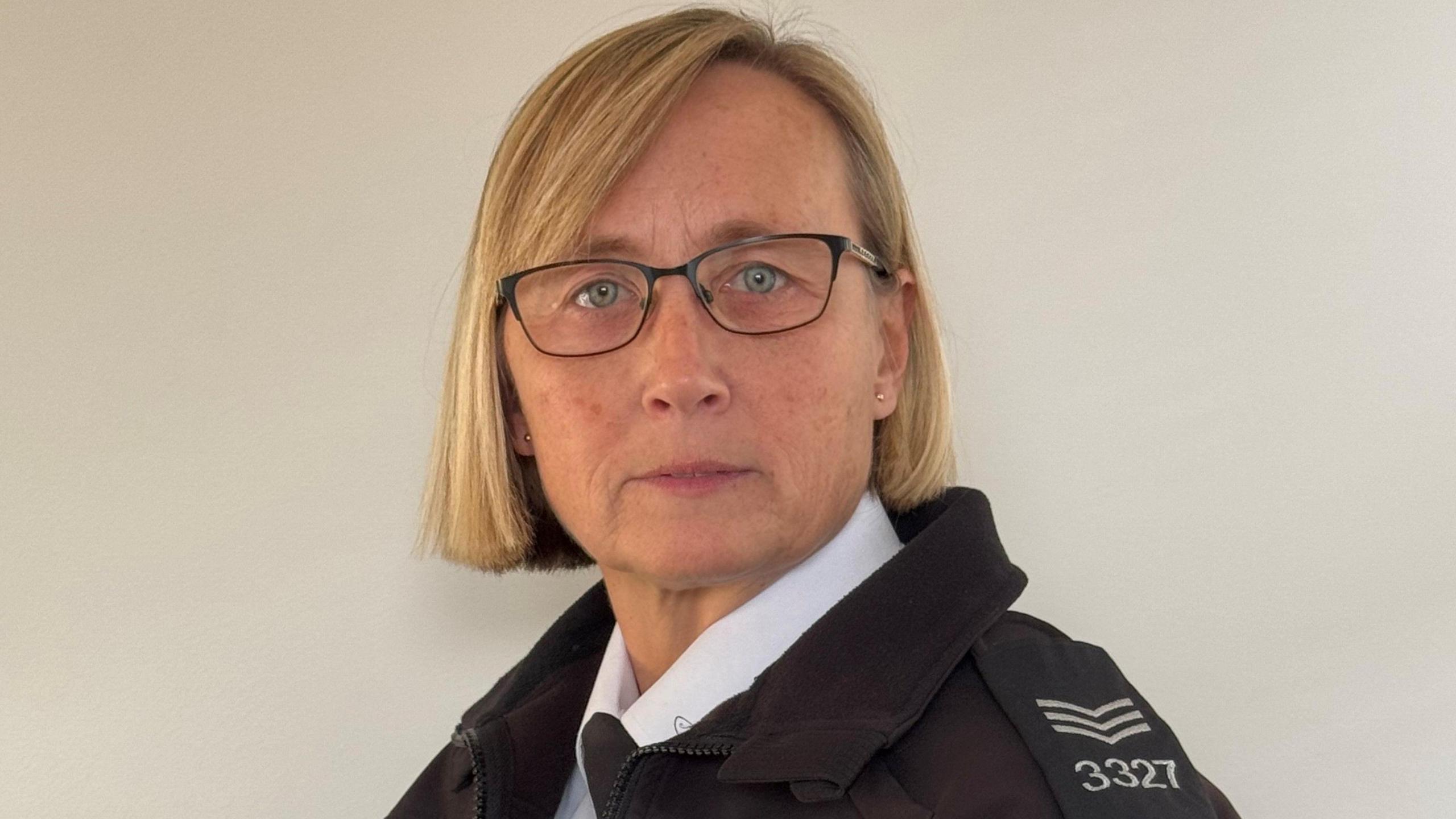 Laura Heggie, who has short blonde hair and wears black rimmed glasses. She is wearing her police uniform, consisting of a white shirt and a black coat that has epaulettes on.