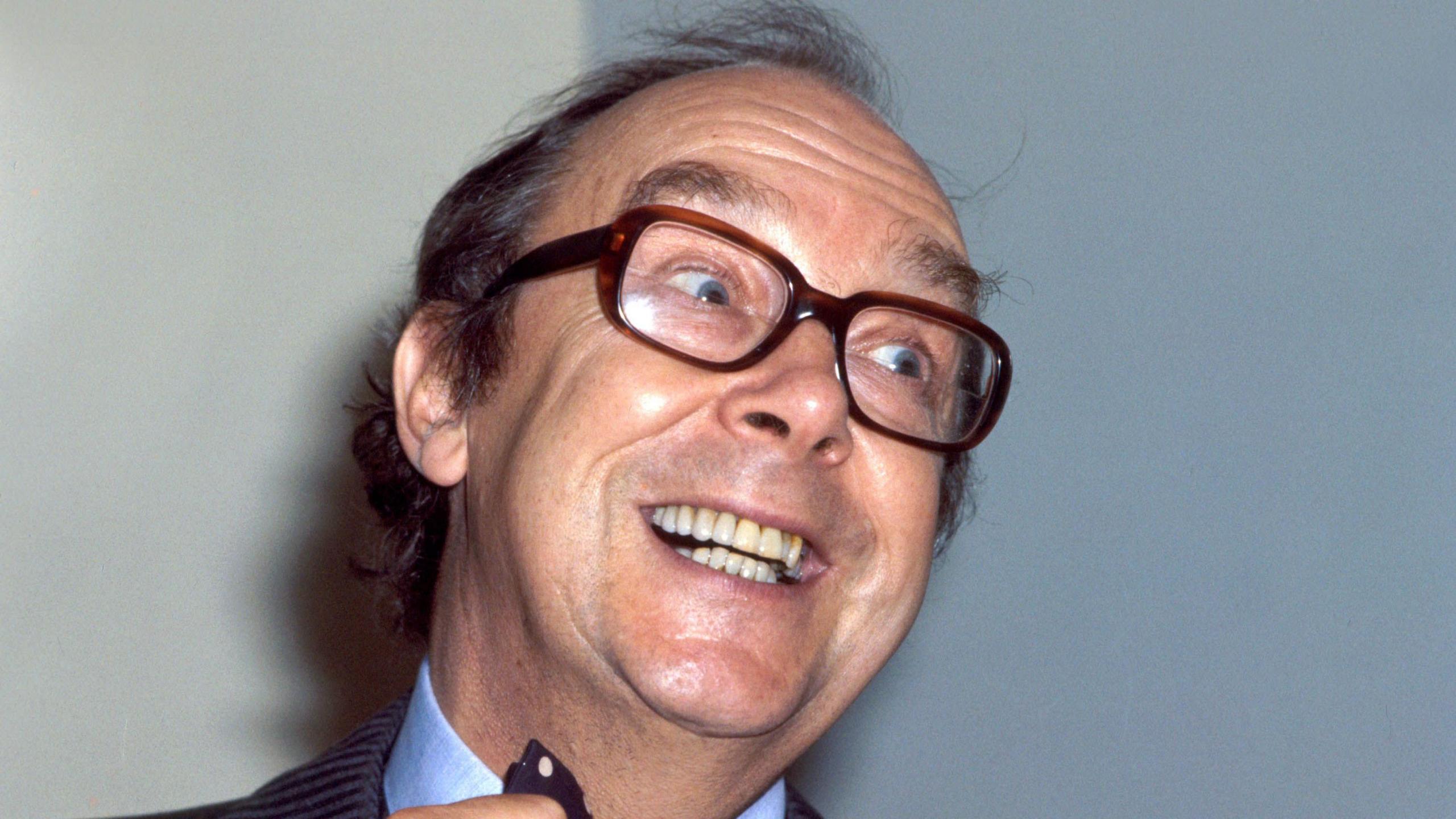 Eric Morecambe looks to the side of the camera. He is smiling and straightening his tie. He wears brown, thick-framed glasses. He has short dark hair.