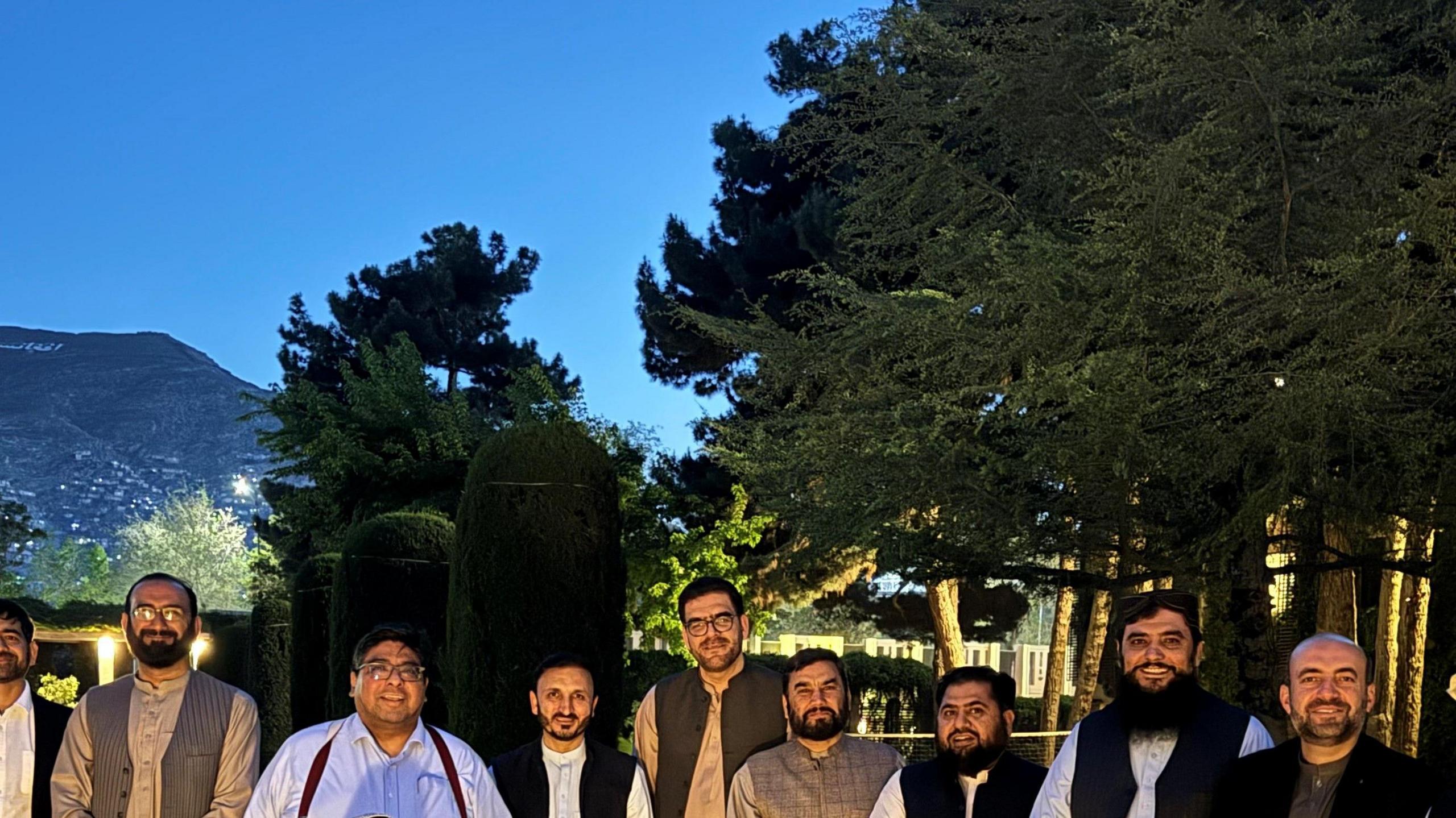 A line of 12 Afghan men, plus Dr Fernando, who is standing behind a woman who is sitting on a chair