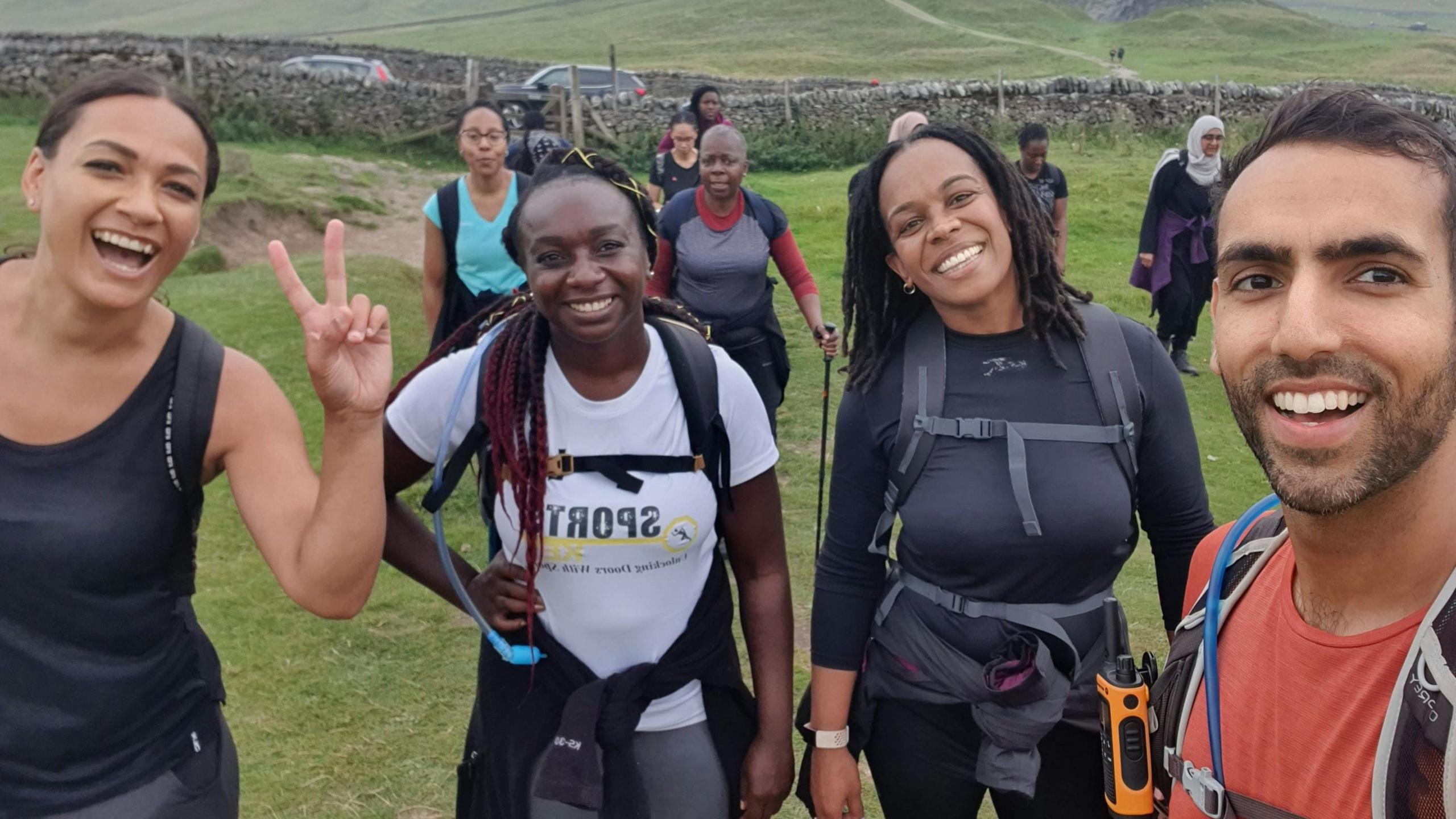 Four people are standing in a field taking a selfie. One woman on the left is holding up her hand in a peace sign. Two women are in the middle with backpacks and walking gear on. Sameed Asghar is on the right, wearing a red t-shirt and a rucksack.