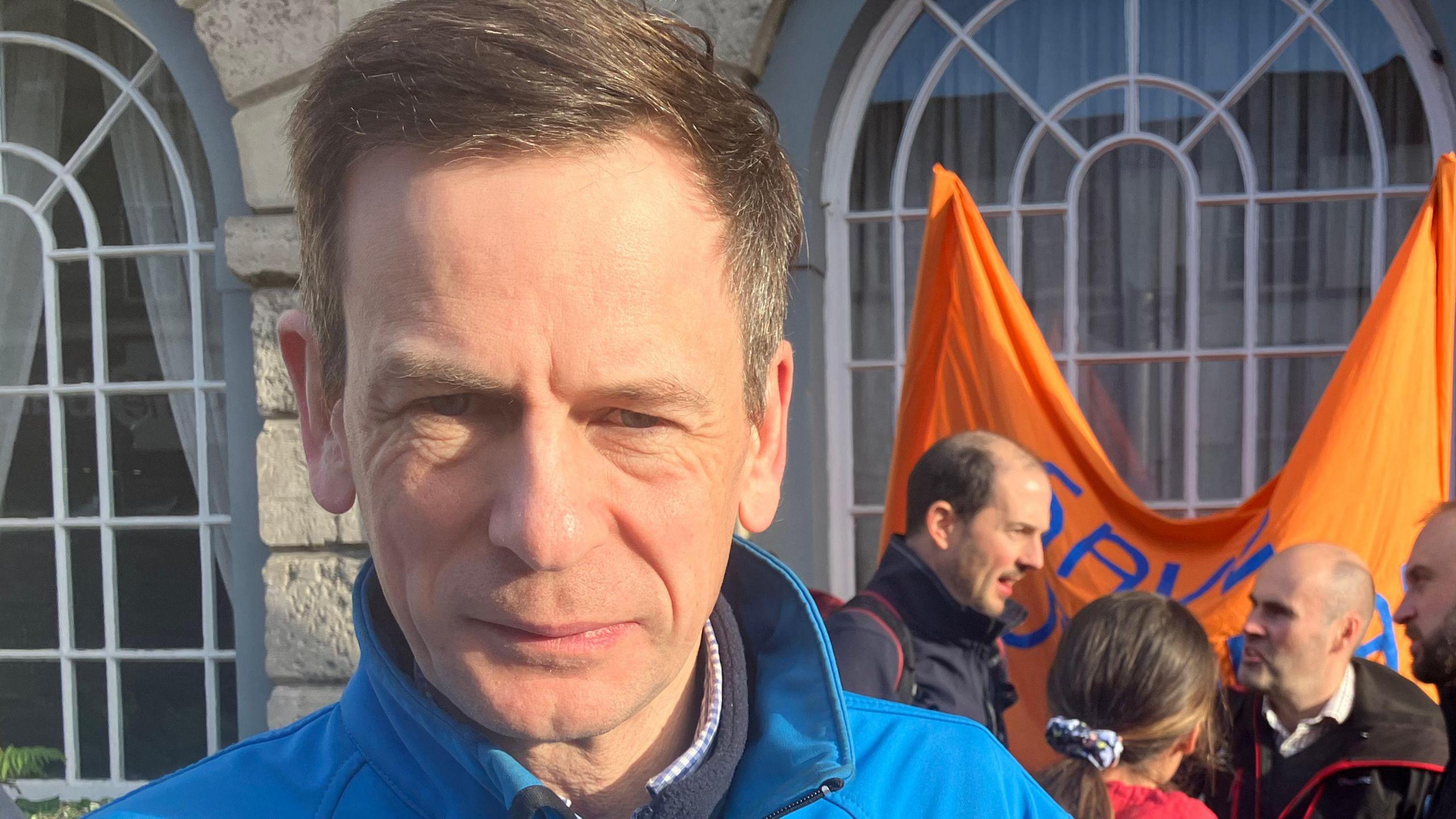 Jeremy Smith, with short greyish brown hair and wearing a blue coat, standing outside of a council building. A group of men and women are chatting behind him. 
