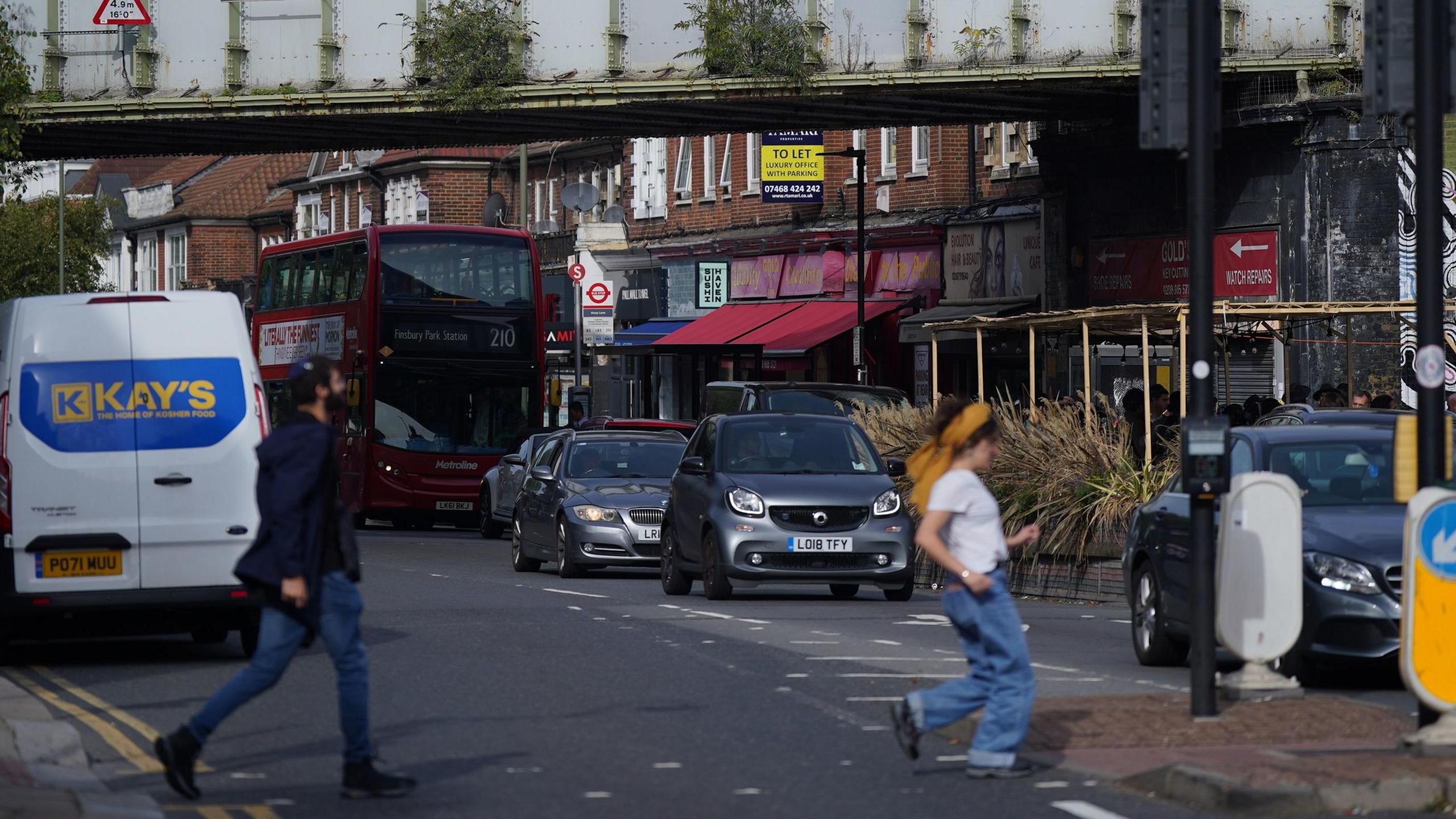 Two people cross a road in Golders Green, with cars, a bus and shops in the background