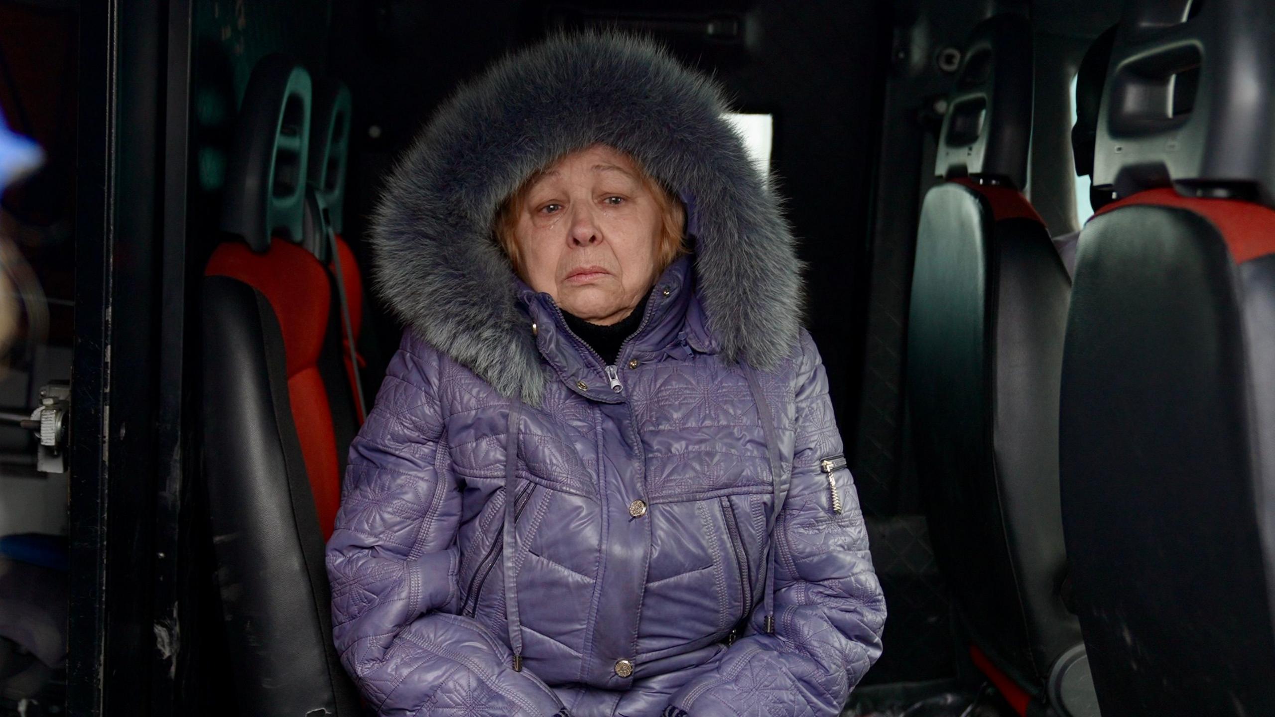 Olga sits inside a van wearing a thick warm purple coat with a fur hood, with a sad and worried expression on her face