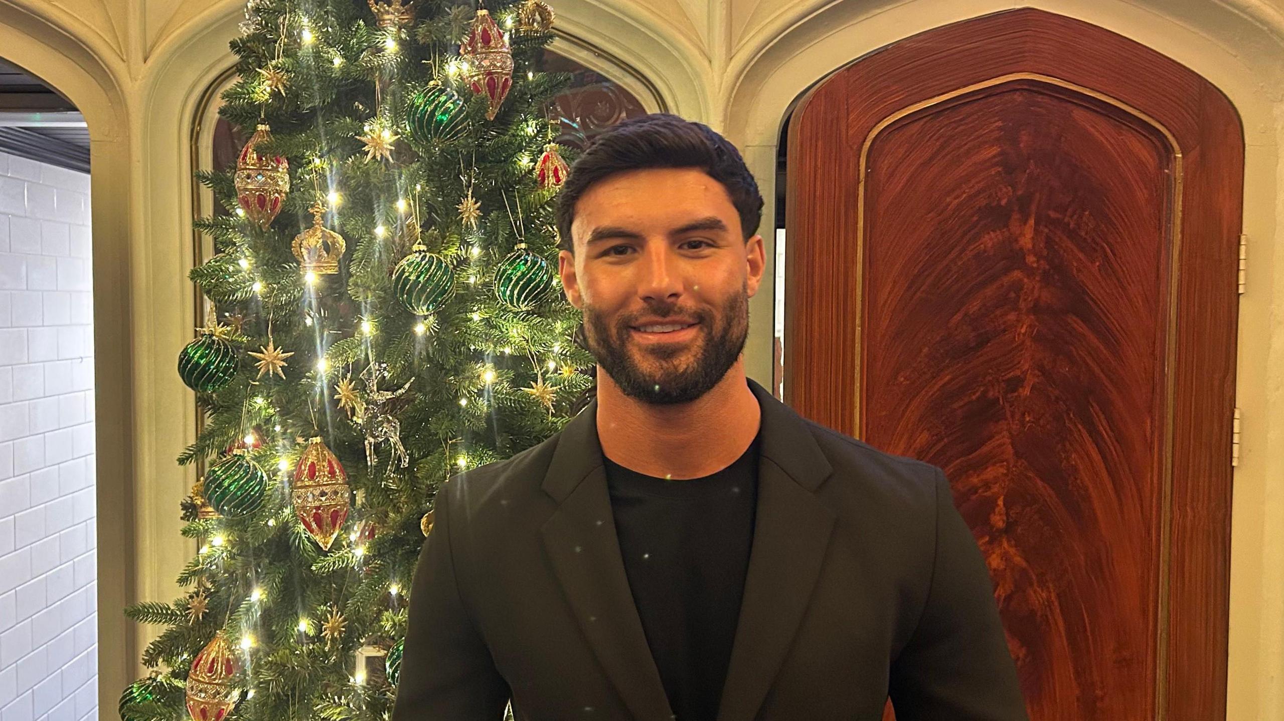 Liam Reardon stands in front of a Christmas tree in a dark suit with black T-shirt.