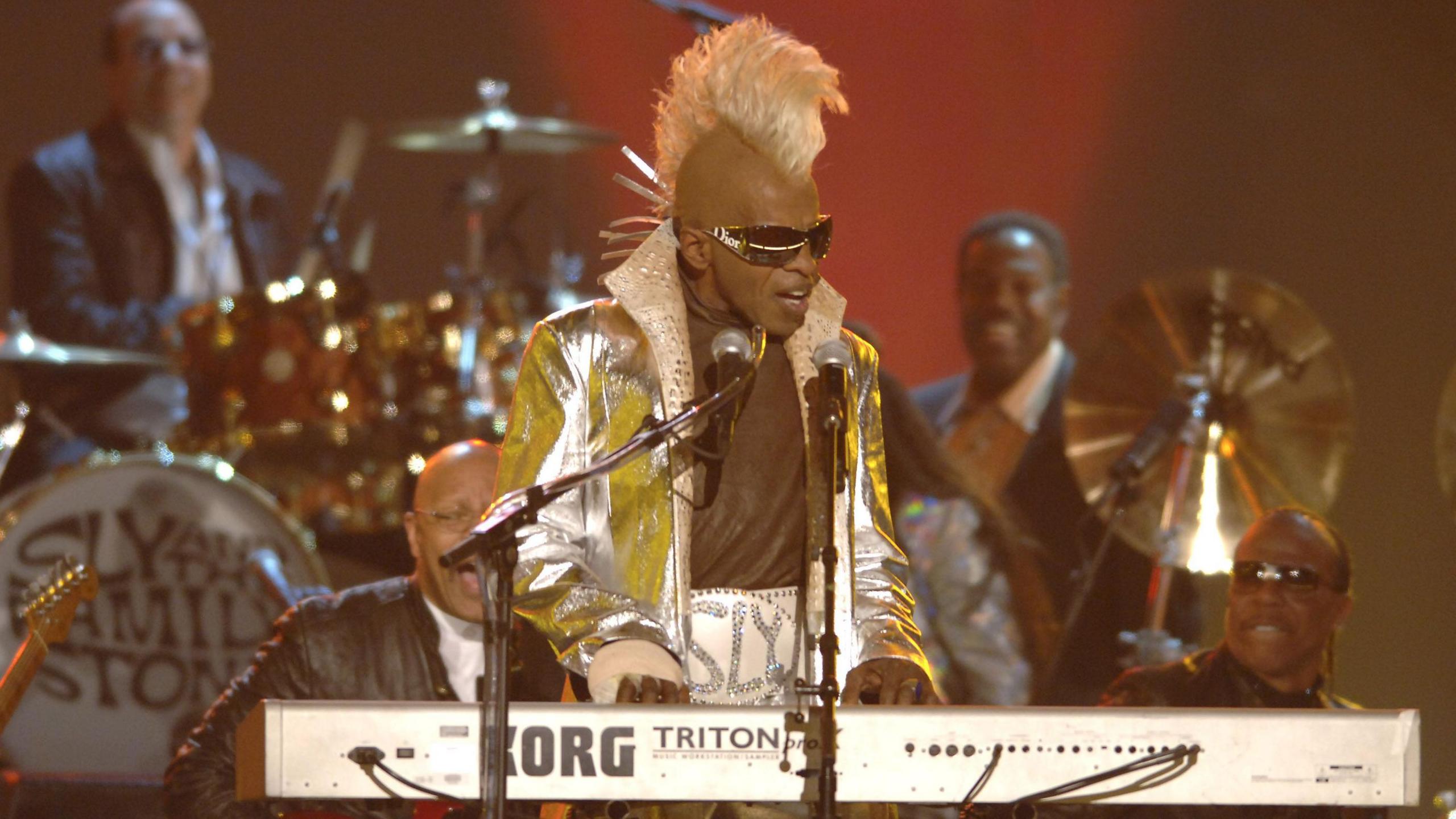 Sly Stone is pictured later in his life wearing a huge white leather belt with the word 'Sly' embellished in silver. He has a matching silver jacket with the collar popped, and a tall bleached mohawk. He plays a white keyboard and sings into two microphones on stands.