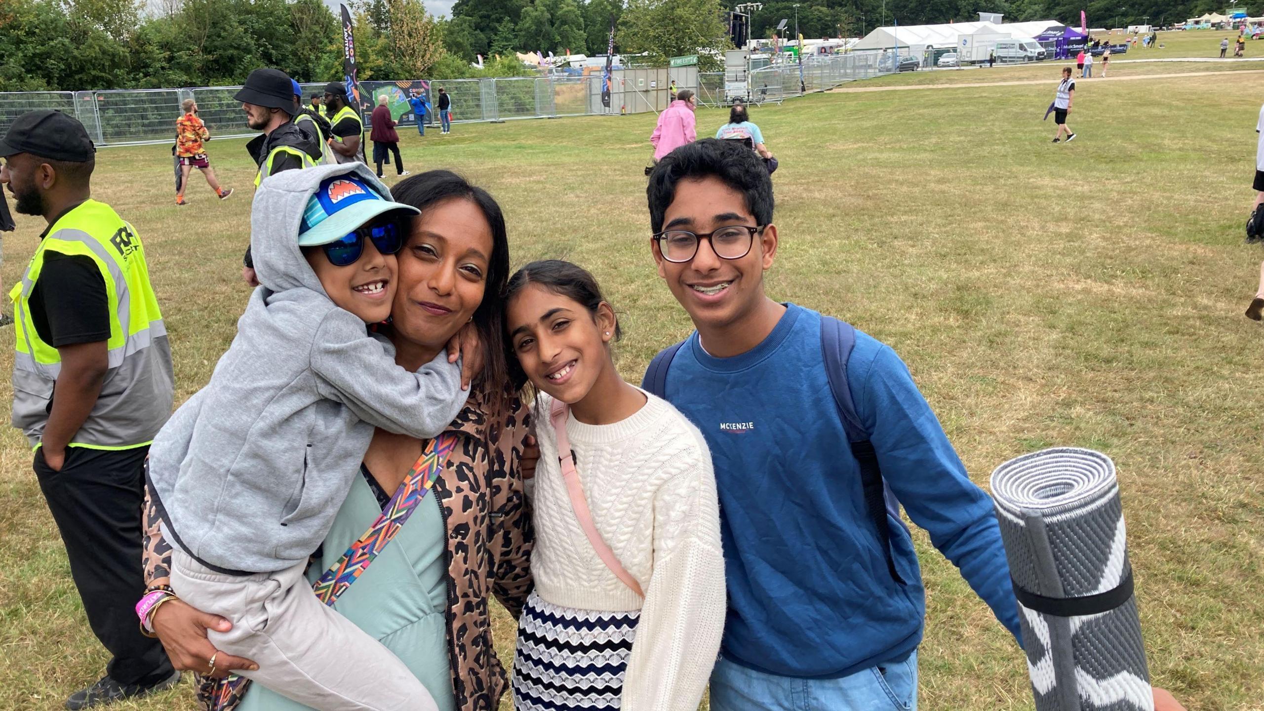 A woman stands with three children in a field. She is holding one of the children, a boy with a grey hoodie. A girl and boy stand next to her. They all have their arms around each other.