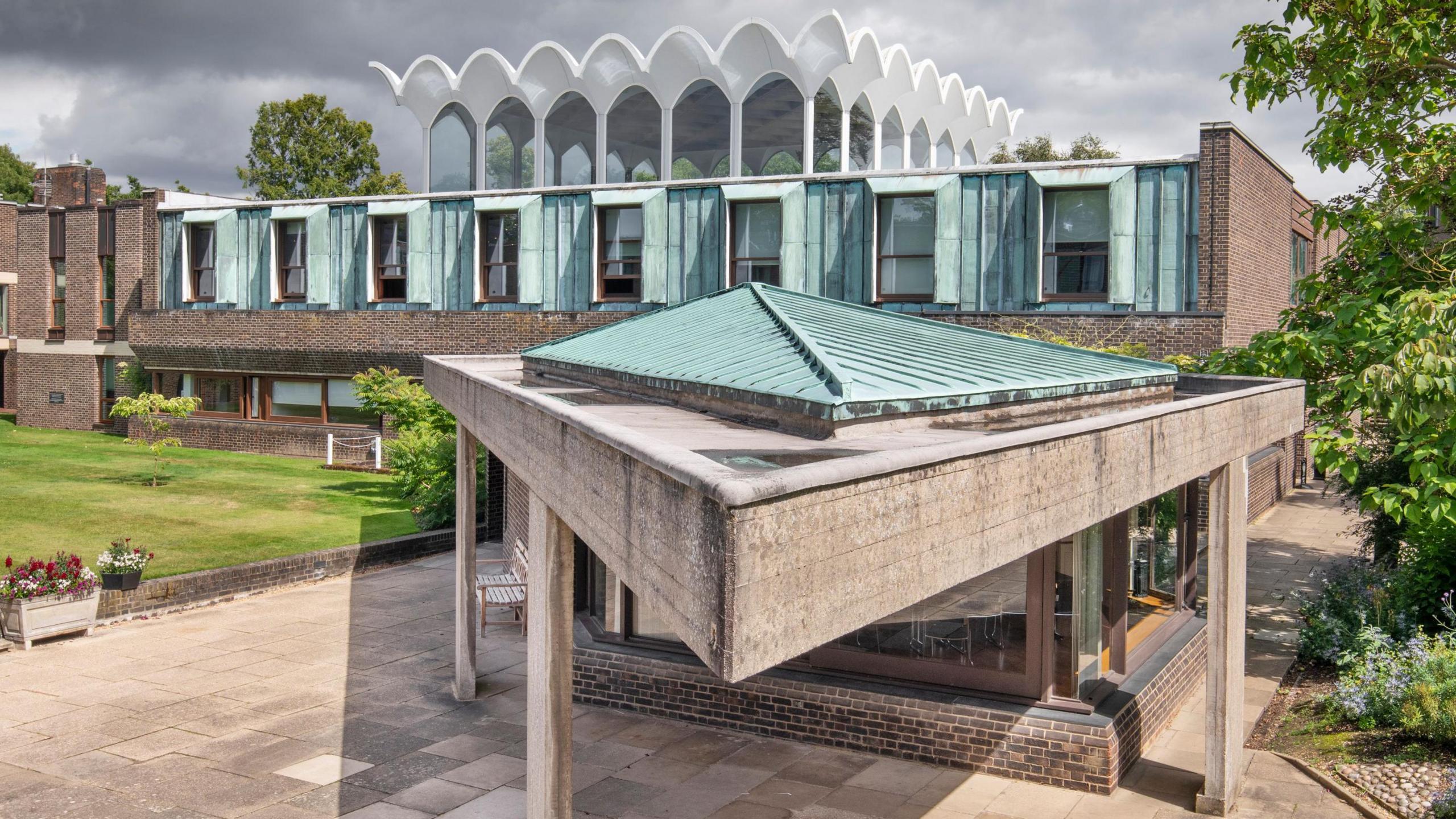 A series of 1960s brick and concrete built, copper roofed buildings with paving to the front, a lawn to the side and above the building at the back an arched roofline, Fitzwilliam College, Cambridge