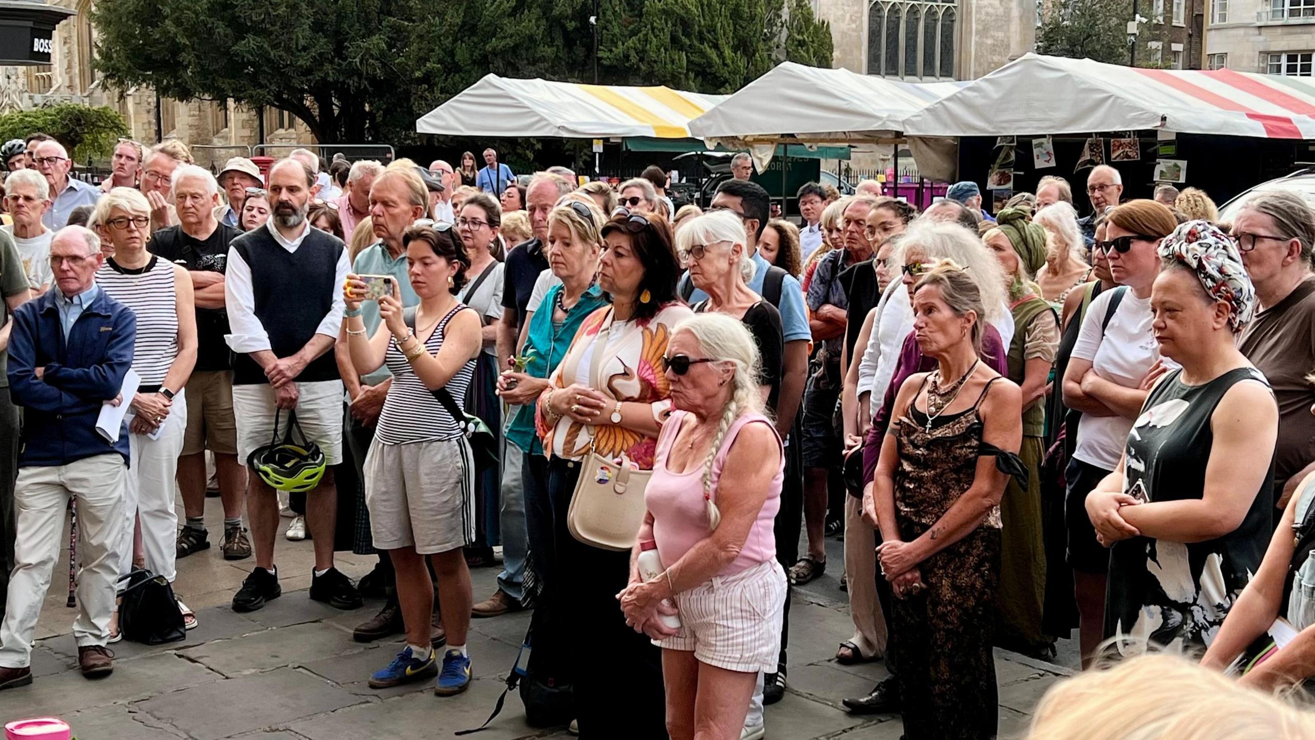 A crowd of people standing in a market square looking on as they are addressed by a speaker at a vigil.
