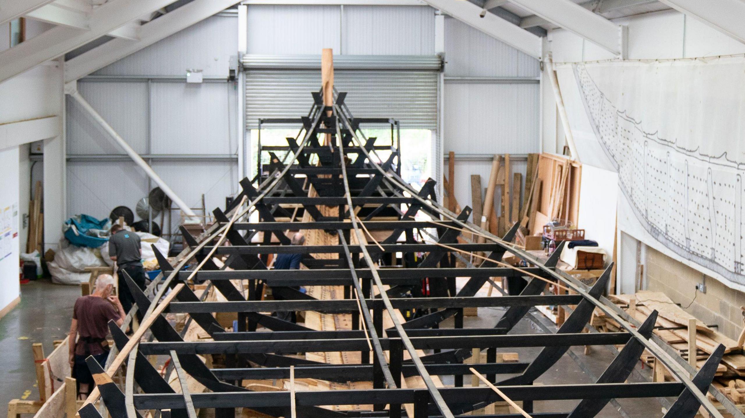 The skeleton of a ship made of wooden panels is pictured inside a large warehouse. People can be seen working on the ship while wooden panels rest all around it.