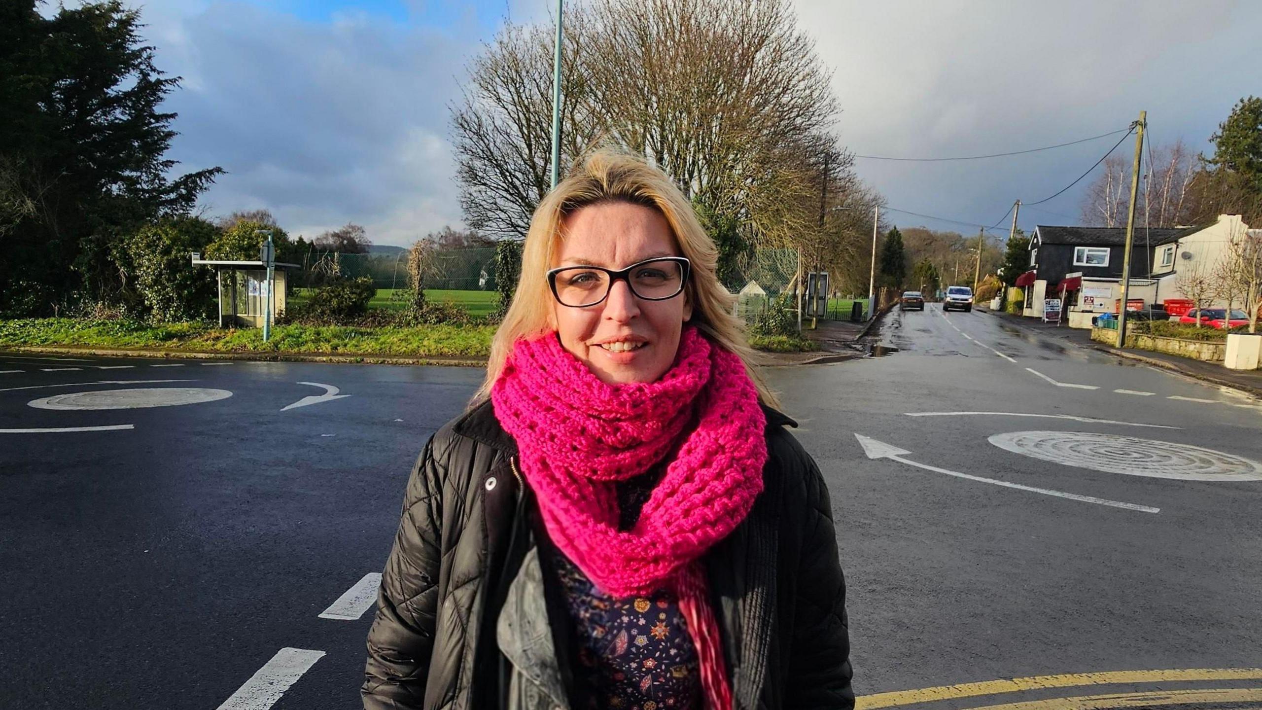 Kristina Walsh is standing by the side of a road in Tutshill. She is wearing a pink scarf and black glasses. Behind her is a roundabout, some residential homes and trees.