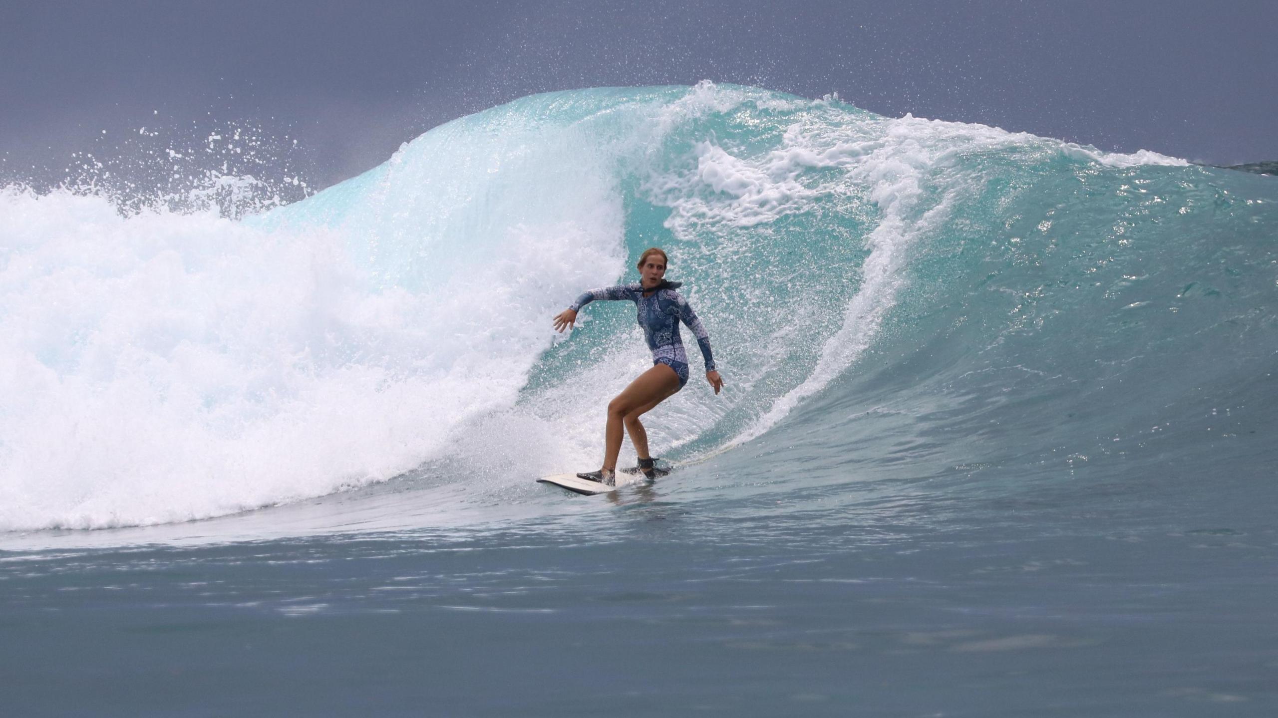 Surfer Dr Ana Manero riding a wave. She is wearing a blue, long-sleeved swimsuit.