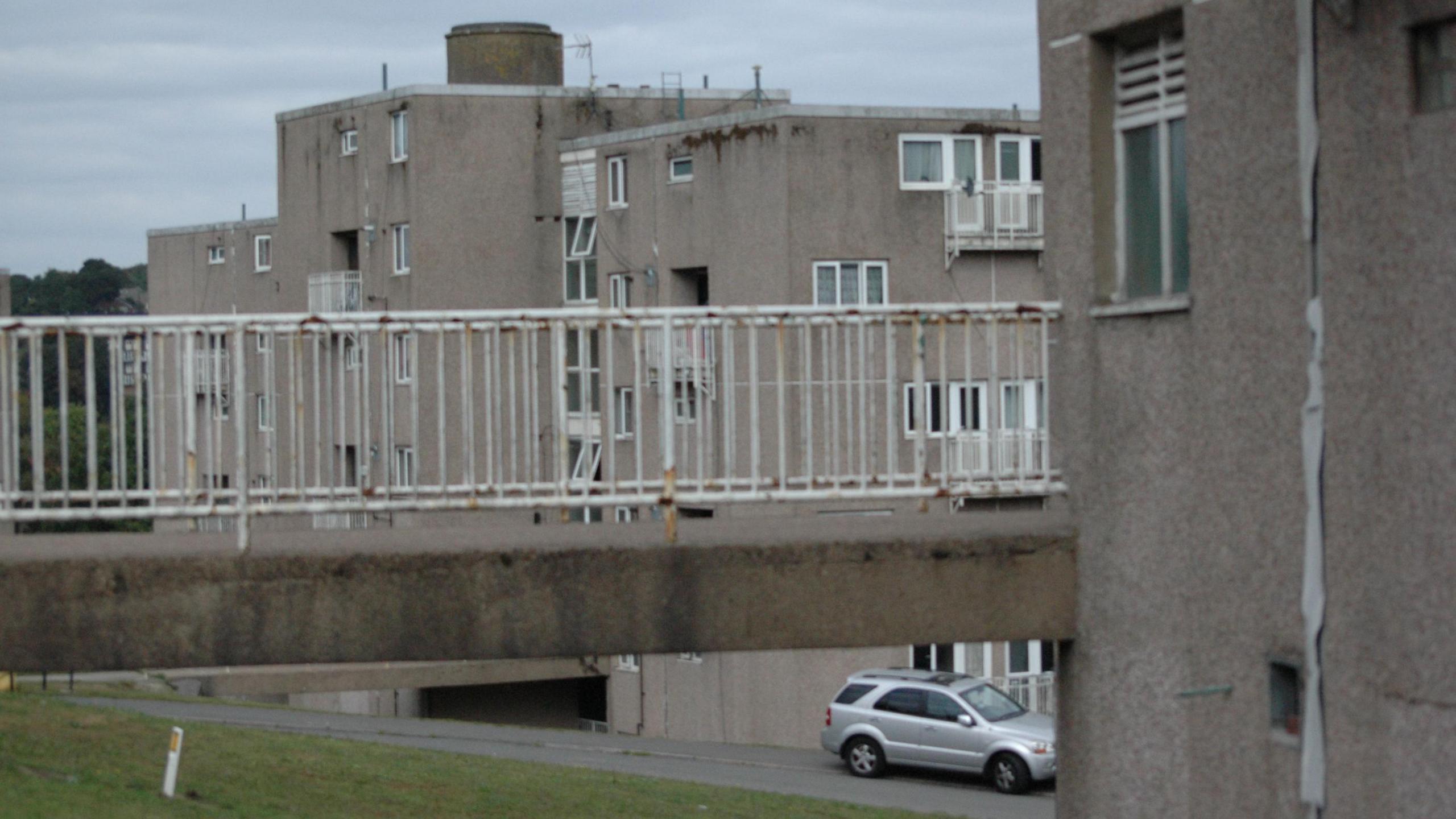 A pebble dash concrete walkway high up above a grass area. There are tired looking tower blocks in the distance.