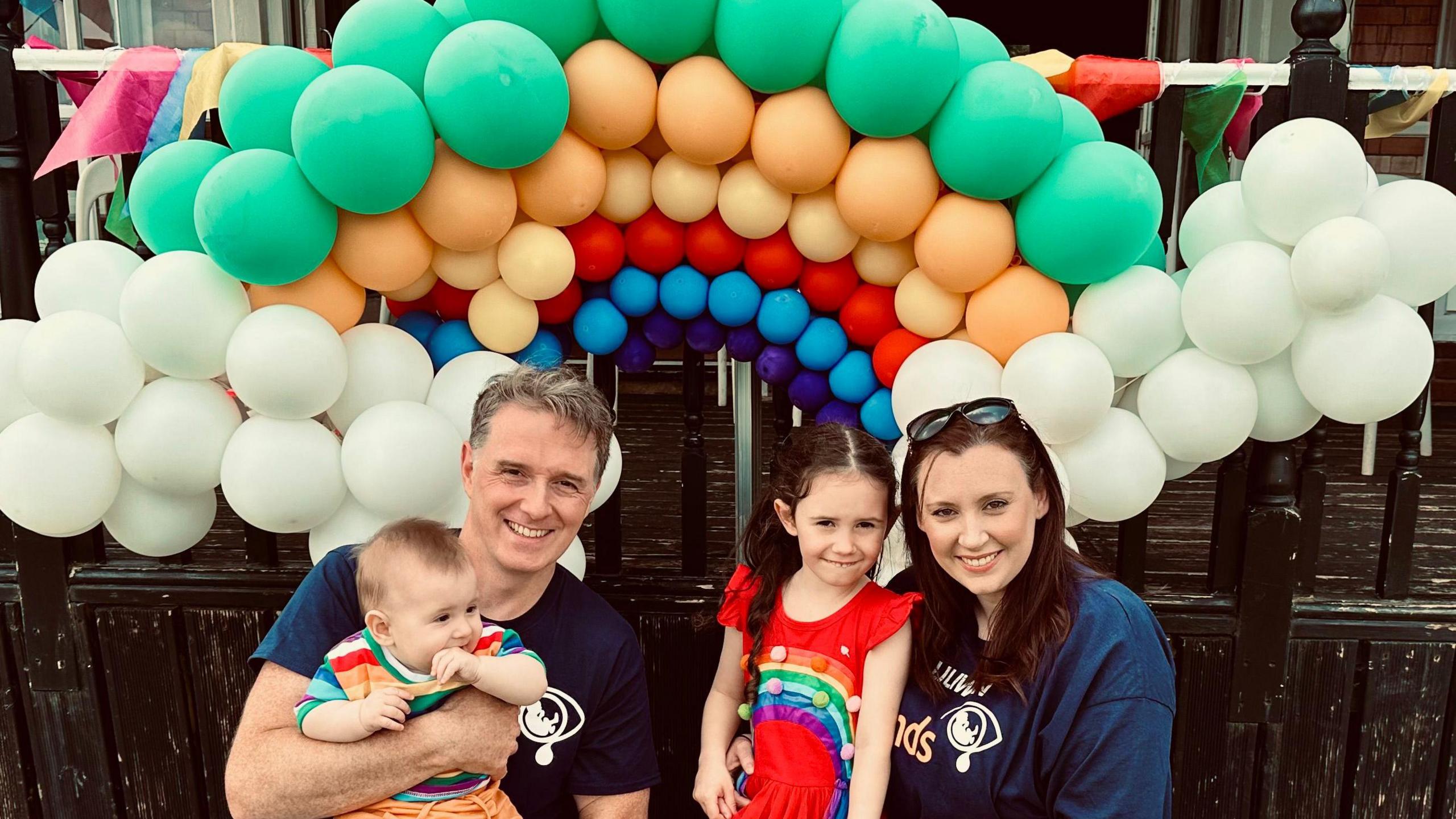 A man with brown hair is holding a baby who is is weaing a rainbow top and orange shorts and chewing his hand.  A woman with brown hair and sunglasses  on her head is next to a young girl with long brown hair who is wearing a red dress with a rainbow on it. They are stood in front of balloons in a rainbow shape.