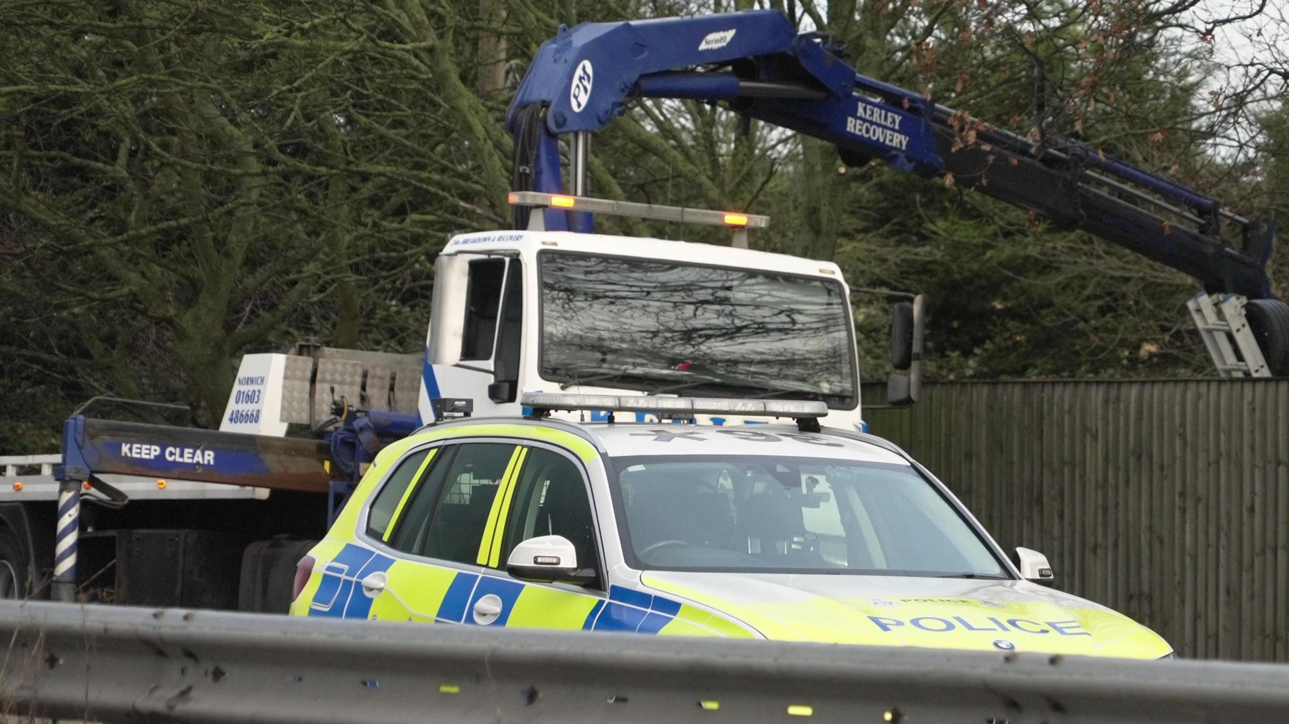 A yellow and blue police car can be seen on a carriageway. Behind it is a white and blue recovery vehicle. 