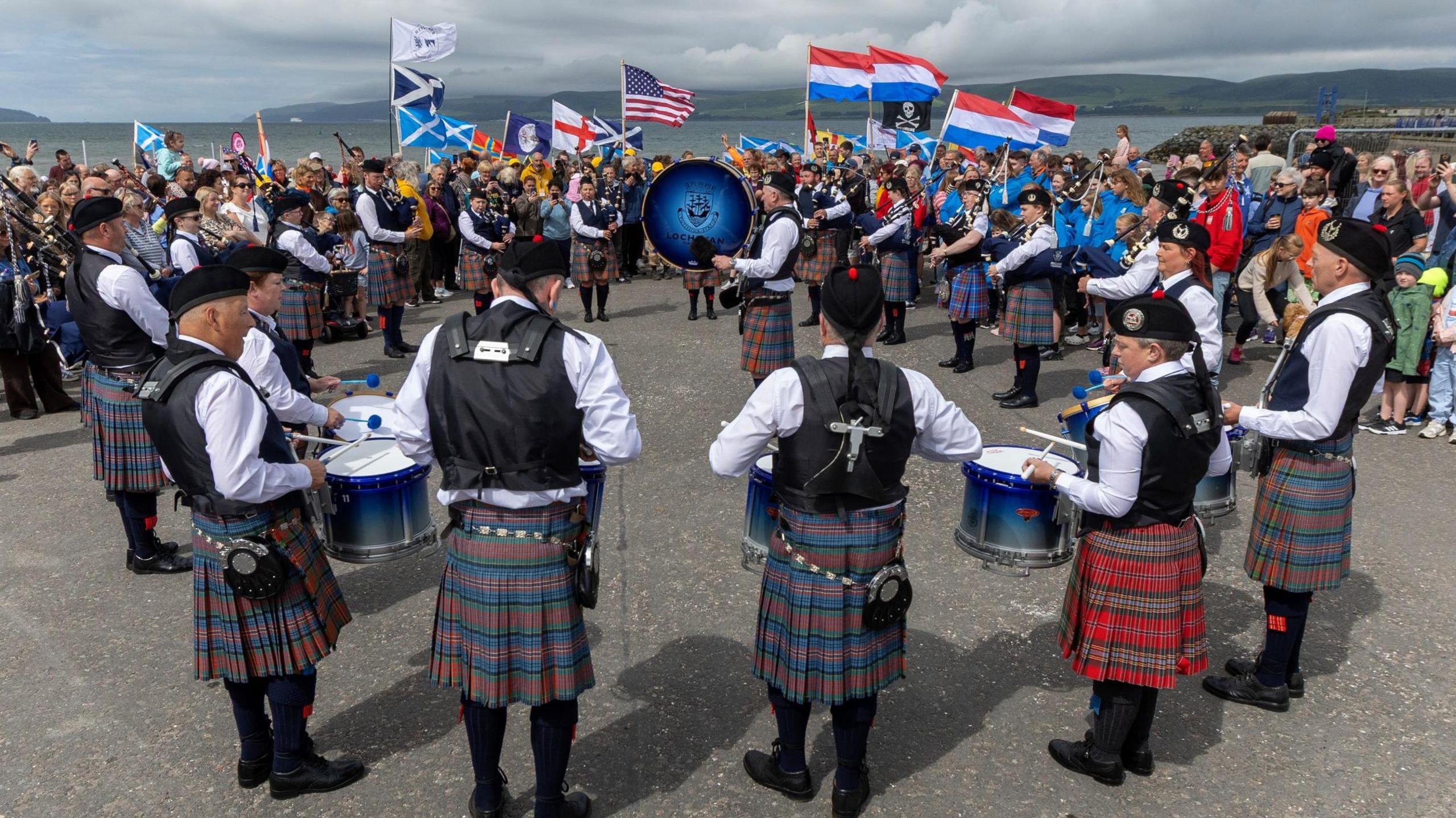 Members of a pipe band standing outside at Straraer harbour. The drummers and pipers are standing in a circle as they preform a tune. A crowd, with some people holding flags of different countries, looks on. The sea and distant shore are in the background.
