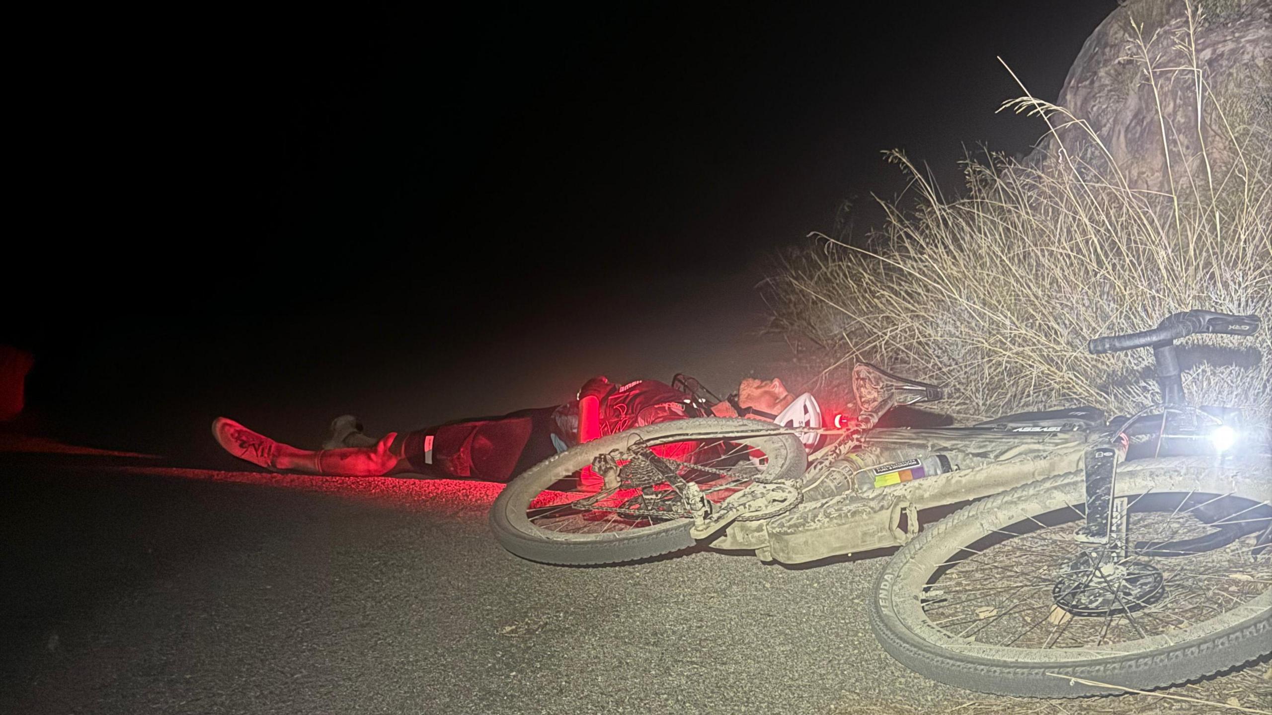 A man wearing cycling gear lying on the ground with his eyes shut, with a bike lying on its side next to him. It's dark and the bike's lights are still on.