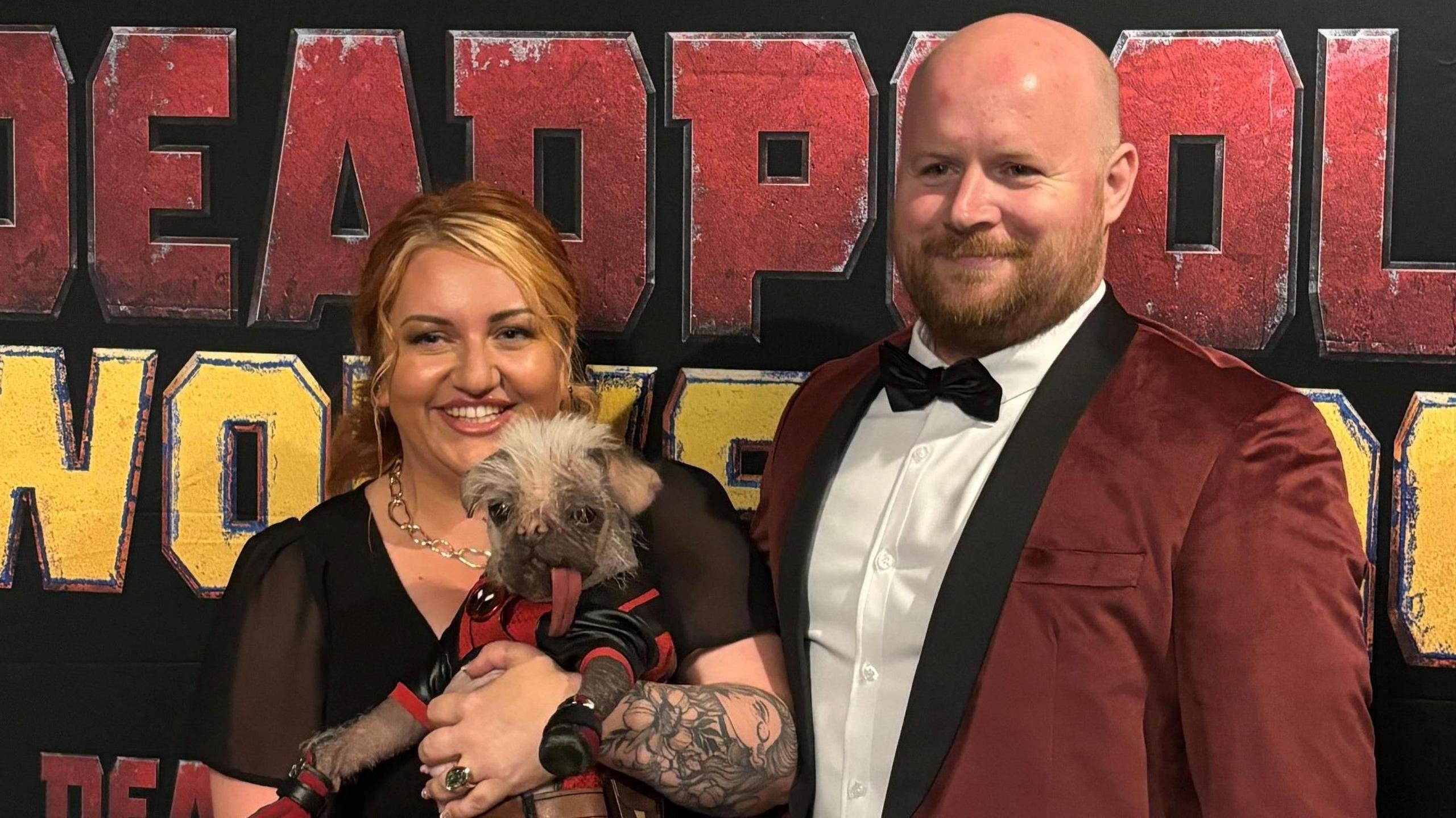 A smiling Holly Middleton, who wears a black dress, gold necklace and has tattoos on her arm, holds her dog Peggy, who is wearing a black and red costume, at an event to promote Deadpool and Wolverine. Her husband, wearing a burgundy dinner jacket, white shirt and black tie, stands next to her.