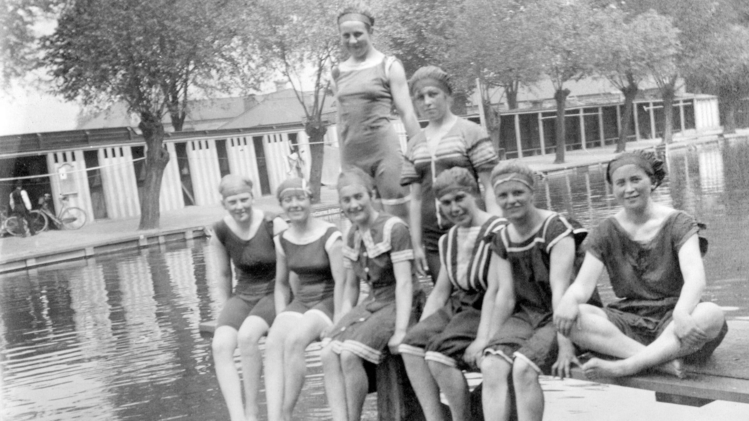 Black and white picture of eight women in swimming costumes sitting beside a river