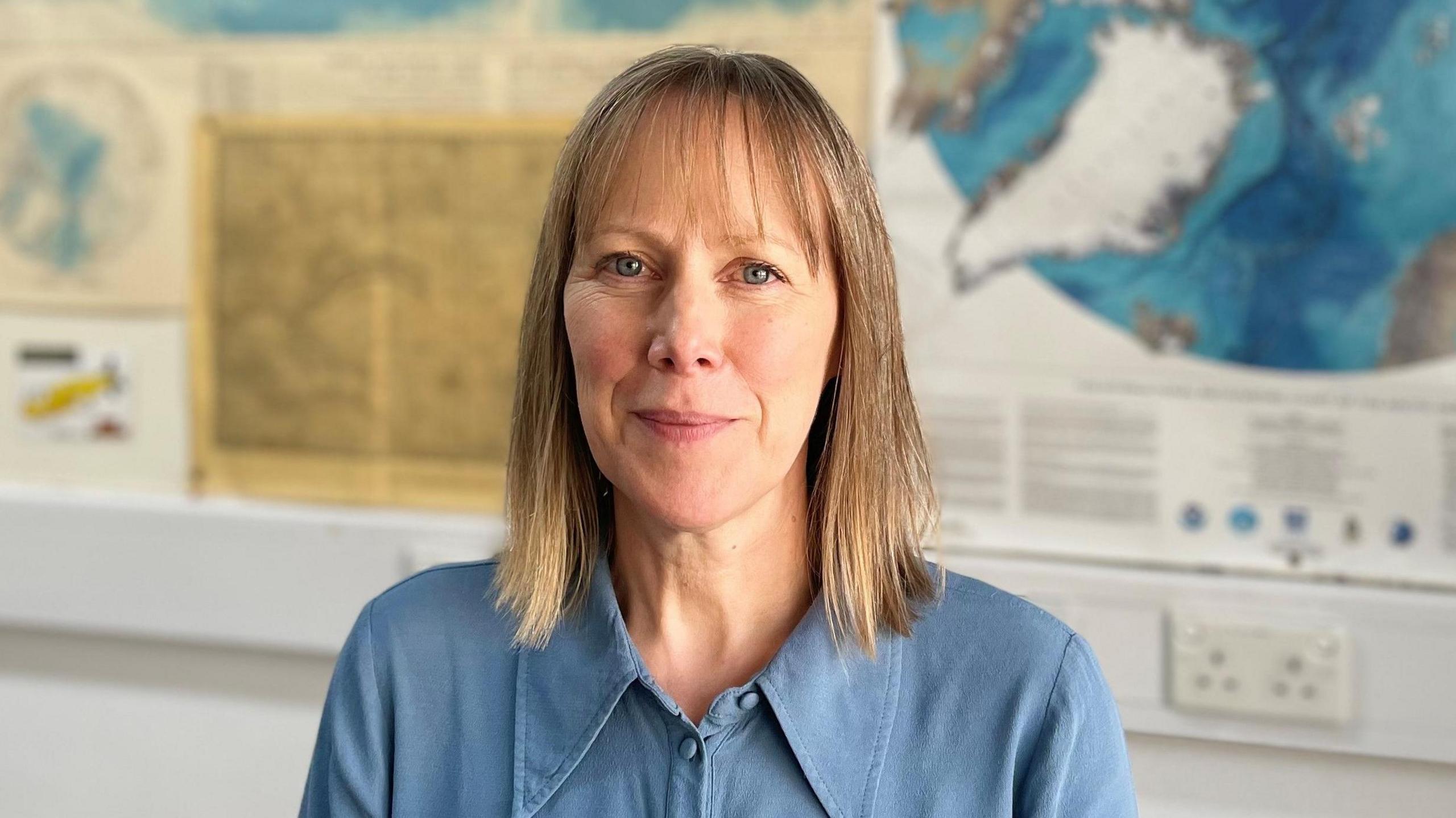 Prof Holliday has medium length brown hair, and is wearing a blue button up blouse. Behind her out of focus are various maps on a wall.