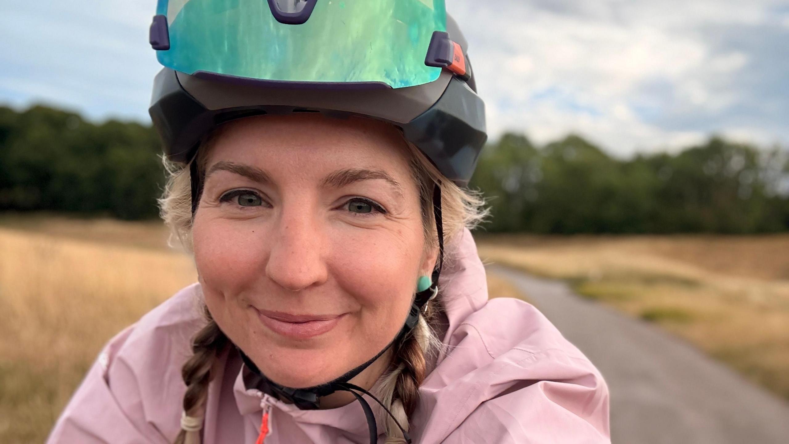 Claire Sharpe wears a cycling helmet and pink jacket has taken a selfie whilst riding her bike through a rural area. She has blonde hair and is smiling. 