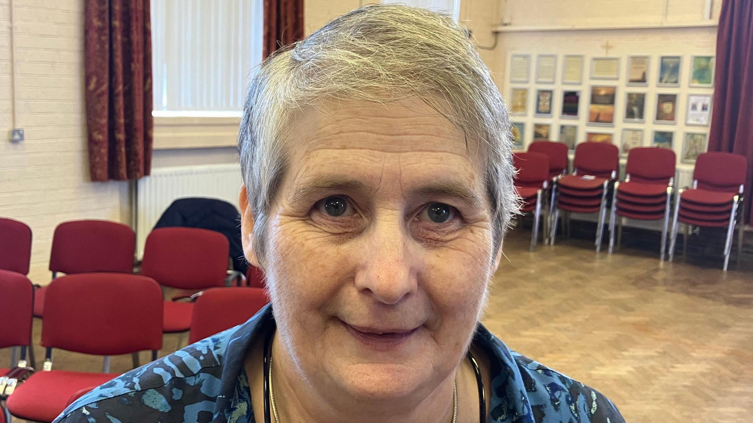 Geraldine McGaughey has short grey hair and is wearing a blue and black top. She's standing in a hall. There are red chairs behind her.