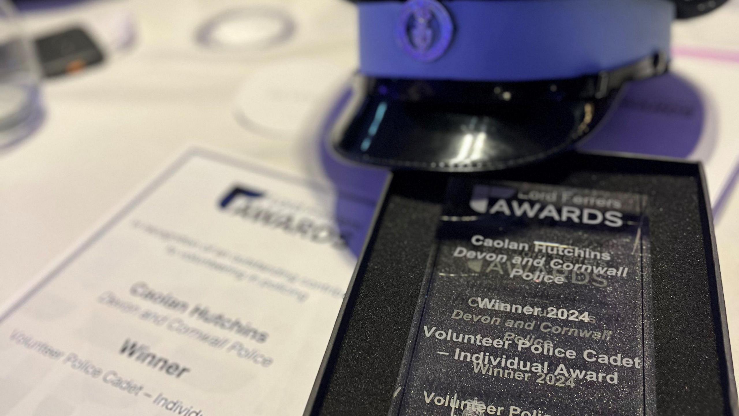 Two awards side by side, made of glass which say "Caolan Hutchins" and "winner" the one of the left is in a case with foam. There is a blue police cap in the back. 