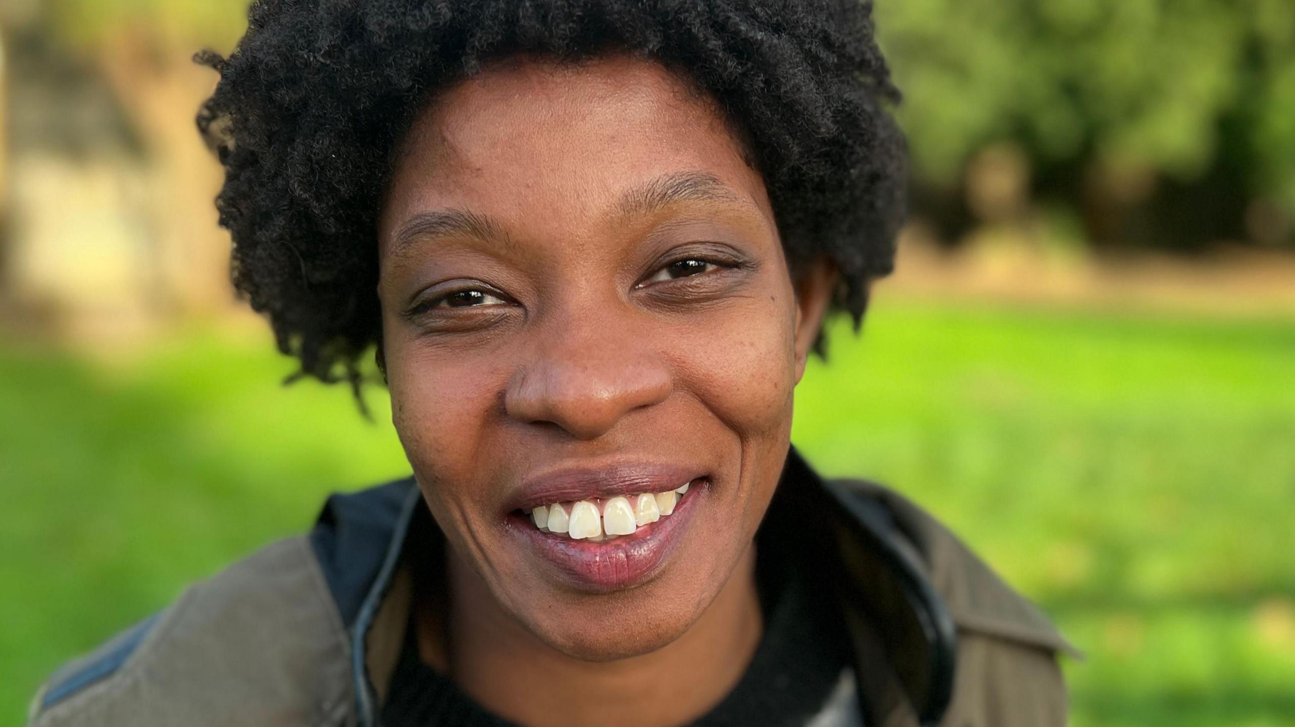 Close up photo of Jessica smiling at the camera. She has brown eyes, black hair and is sat outside during the day. She has a khaki green coat on and stripy jumper. Behind her is a patch of bright green grass, highlighted by a patch of sunlight.