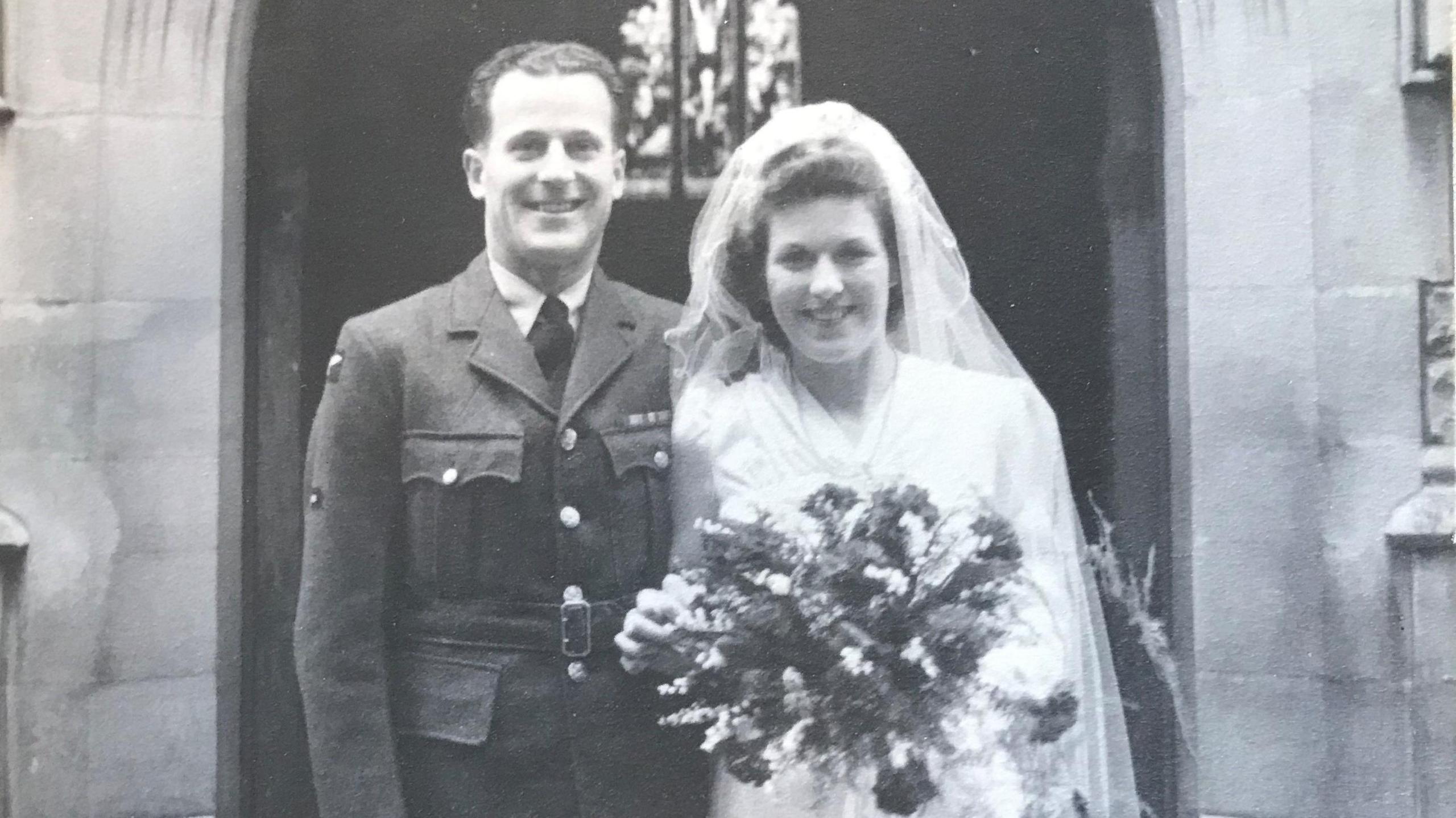 A black and white photograph of Mrs Bond and her husband stood outside a church on their wedding day. Hilda is in a traditional white bridal dress holding a bouquet of flowers and her husband is wearing military uniform