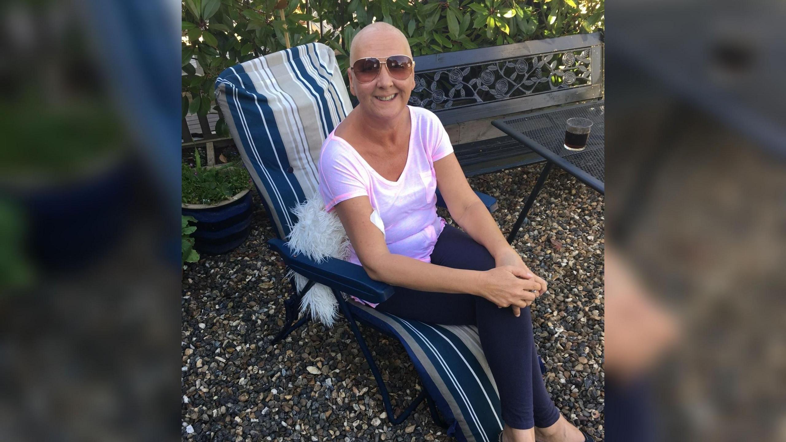 Dawn Willis sits on a cushioned garden chair and looks at the camera. She is wearing sunglasses and smiling at the camera. She is wearing a purple t-shirt and has a bald head.