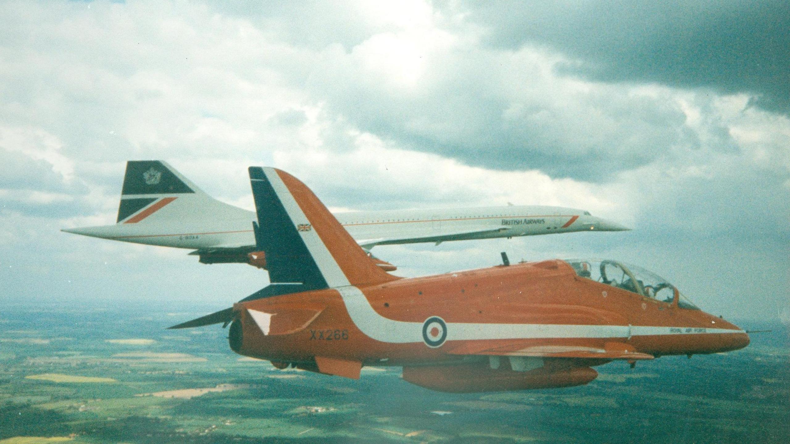 A classic Red Arrows plane flies alongside a Concorde passenger jet high above the ground. Fields can be seen below.