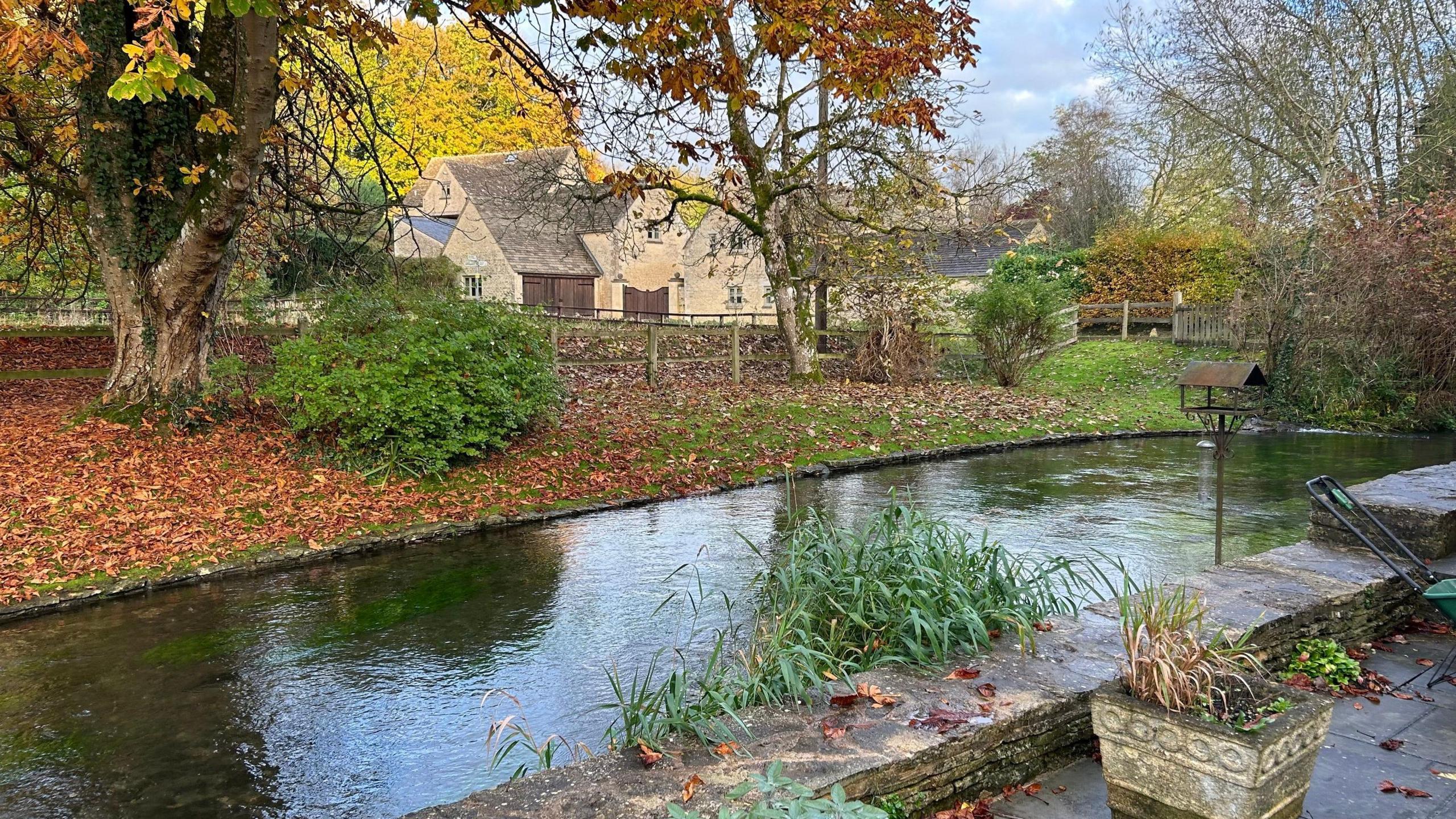 The River Coln in the Cotswolds, pictured from someone's back garden. A short Cotswold stone wall borders it, and on the opposite bank, there is grass, trees, and a fence which borders the garden of a Cotswold stone house that can be seen.