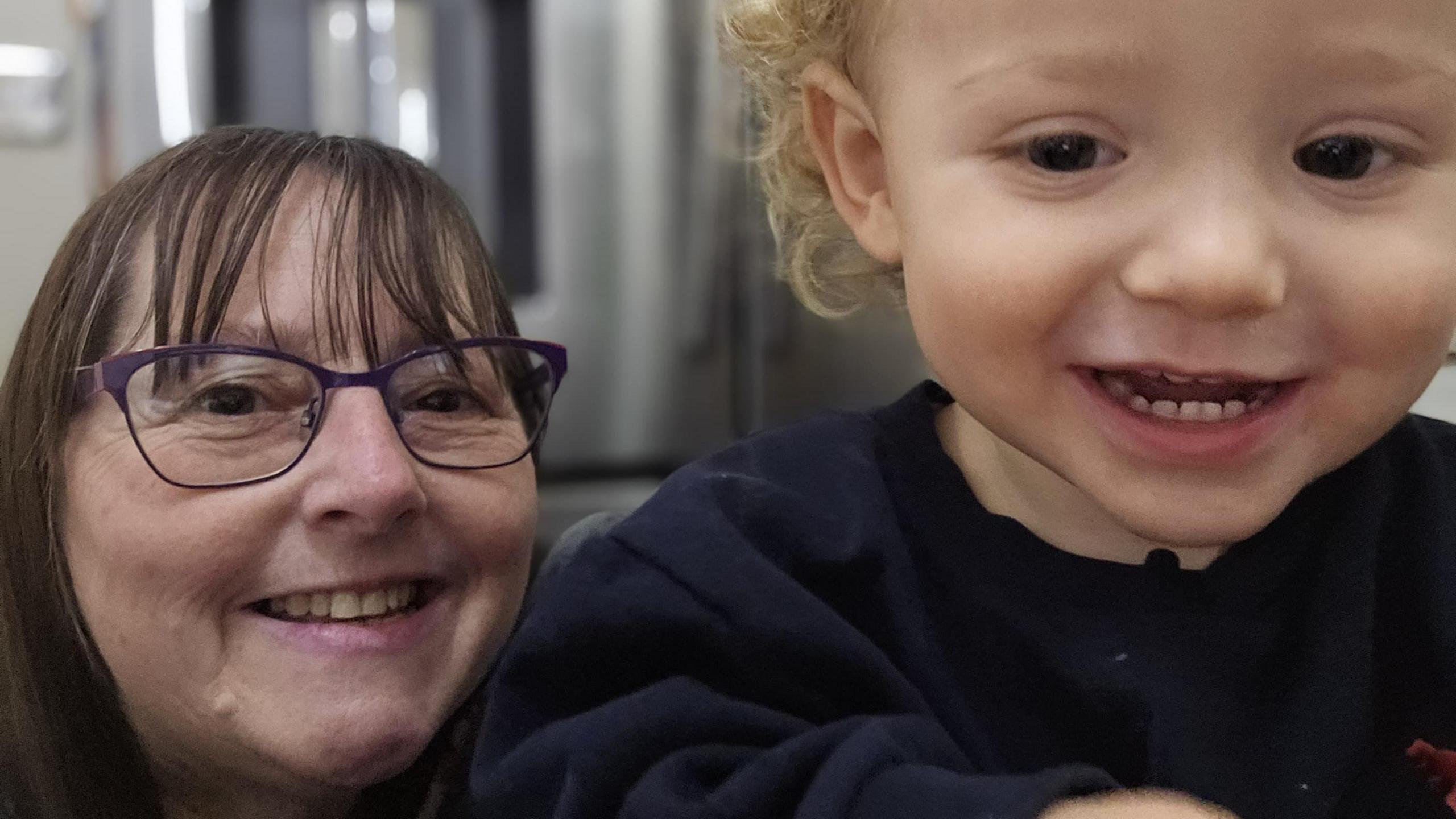 Judy, who is wearing glasses and has brown hair with a fringe takes a selfie with her grandson who has blonde curly hair and is smiling at the camera.